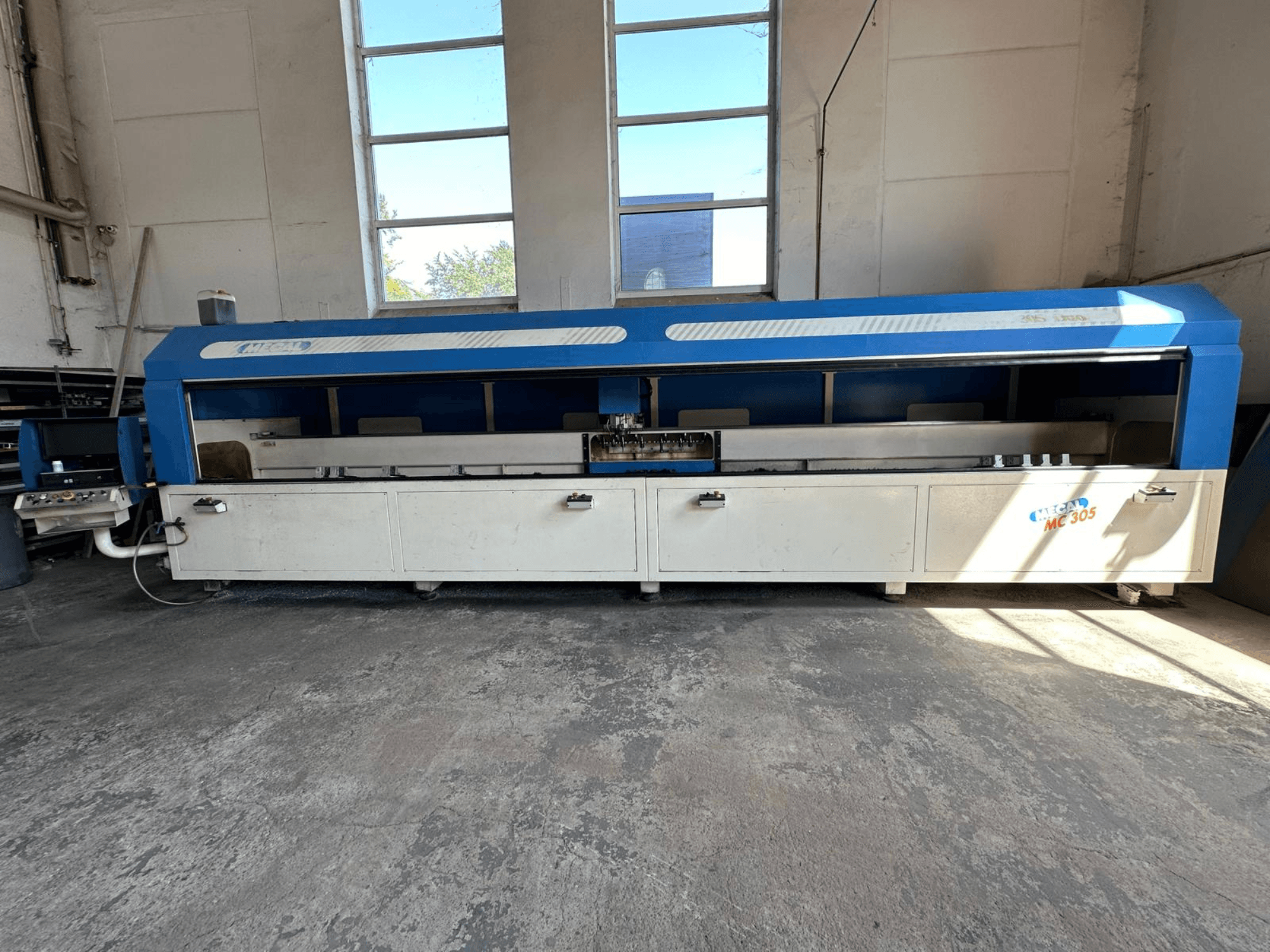 Вид спереди вертикального обрабатывающего центра MECAL MC 305, выполненного в сине-белом цвете, с видимым рабочим столом и панелью управления.