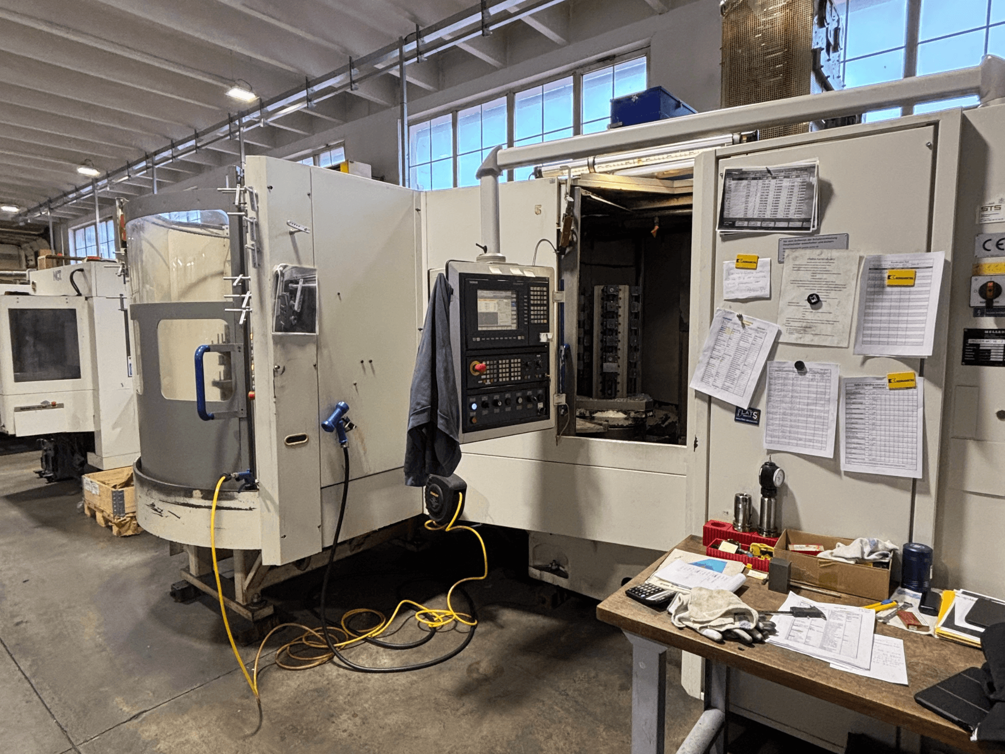 Станок с ЧПУ в мастерской, вид спереди, панель управления и документация сбоку; инструменты и материалы на столе.