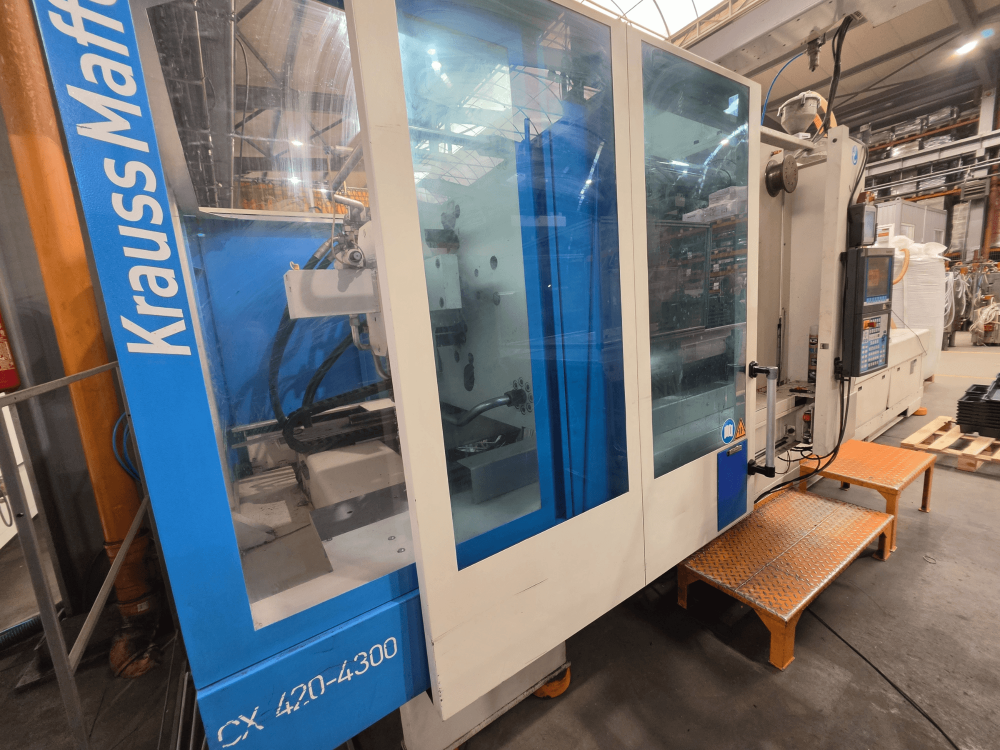 Гидравлическая машина для литья под давлением Krauss Maffei CX 420-4300, вид сбоку, видна панель управления и прозрачные панели.