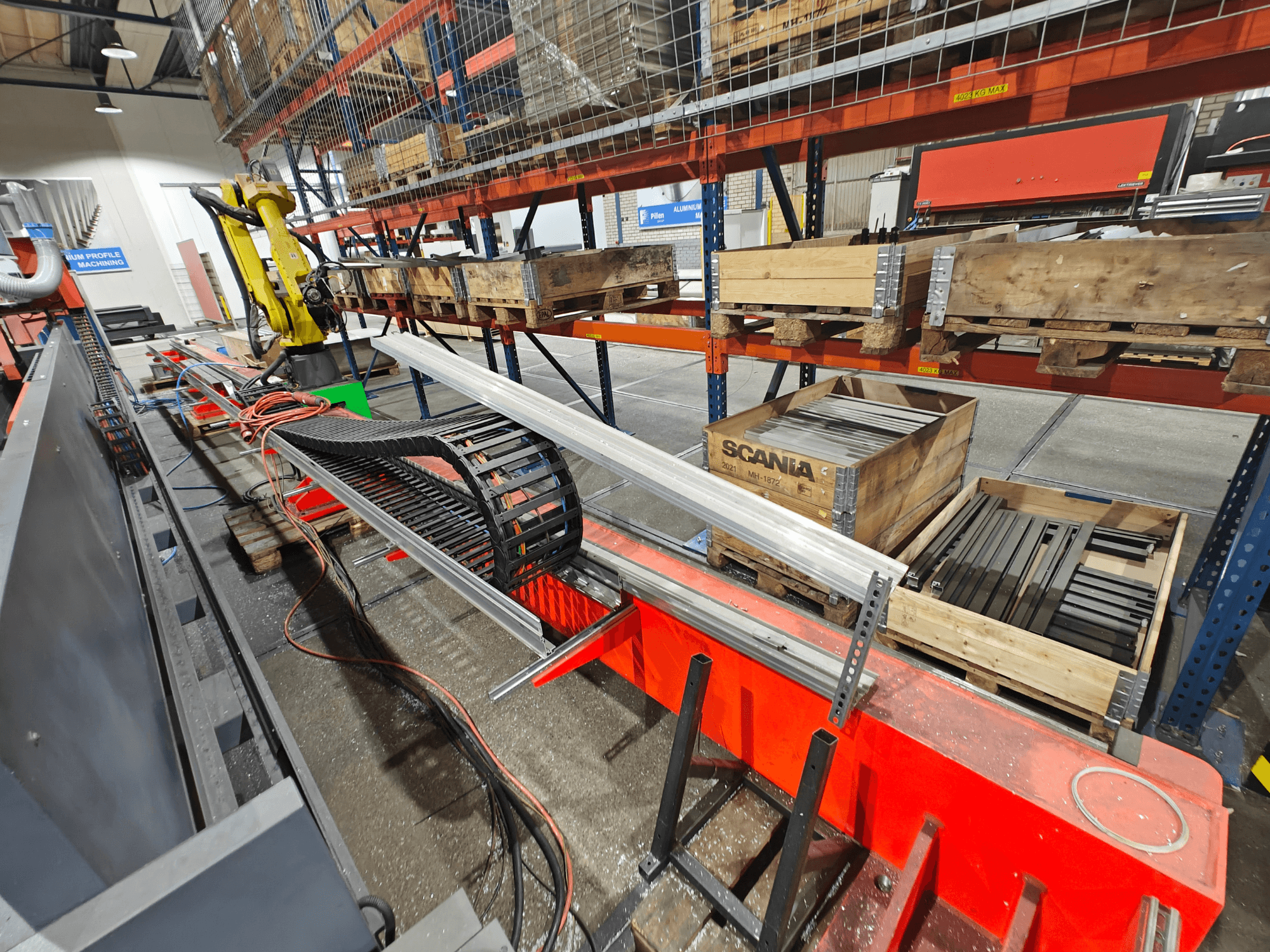 Роботизированная рука на рабочей станции, видны оранжевые и черные кабели, ящики Scania и алюминиевые профили в заводских условиях.