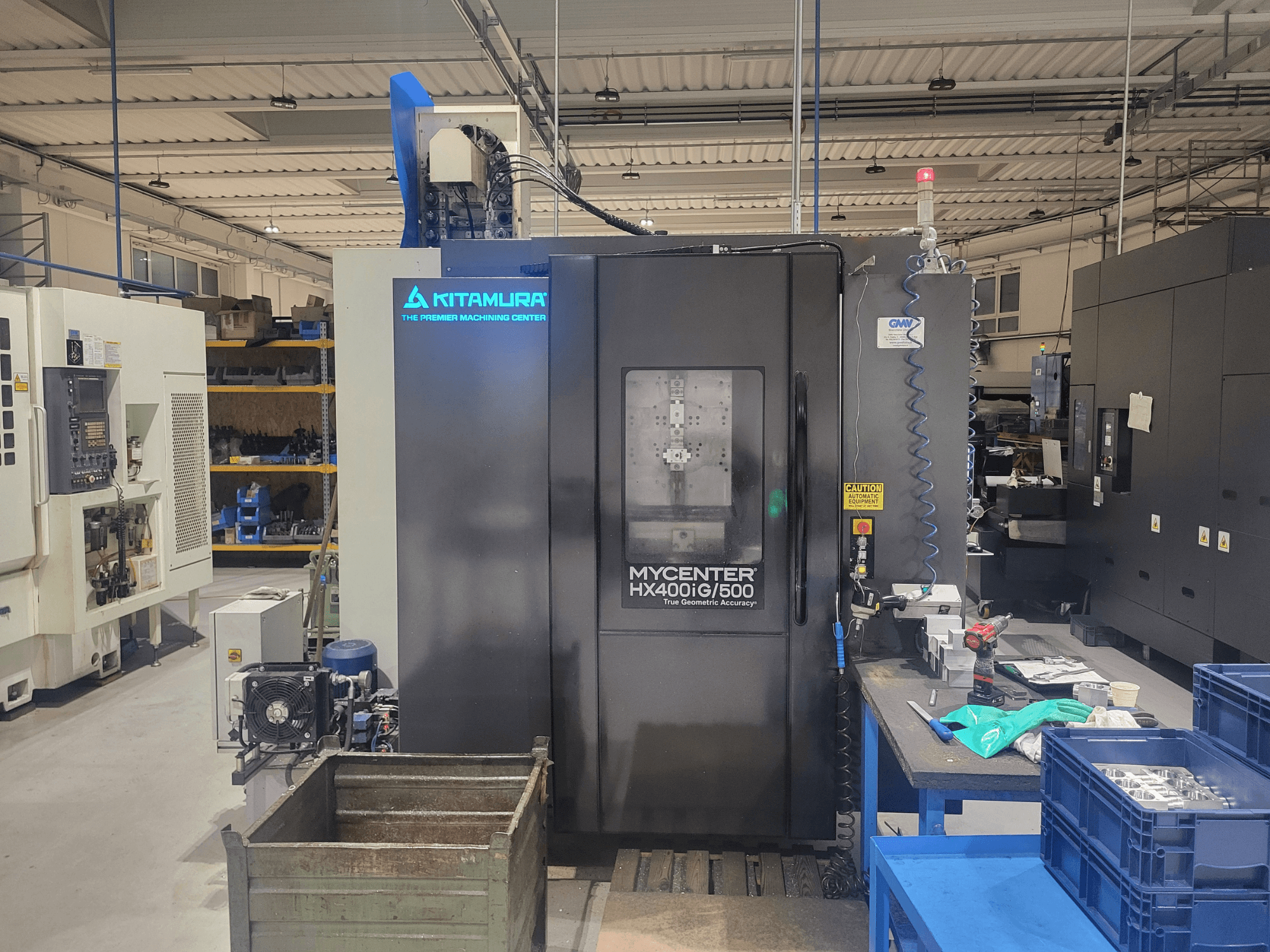 Станок с ЧПУ Kitamura Mycenter HX400iG/500 в мастерской, вид спереди, с элементами управления, складом инструментов и материалов рядом.