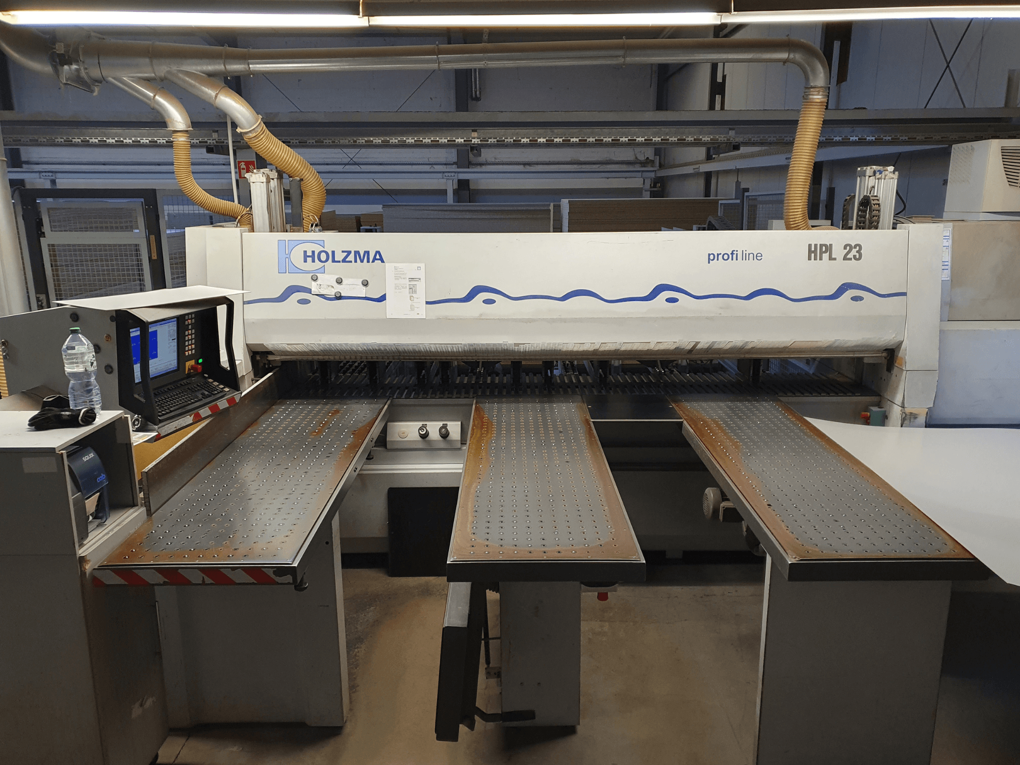 Резальная машина HOLZMA HPL 23, показанная спереди, с тремя перфорированными столами, компьютерным терминалом и бутылкой воды рядом.