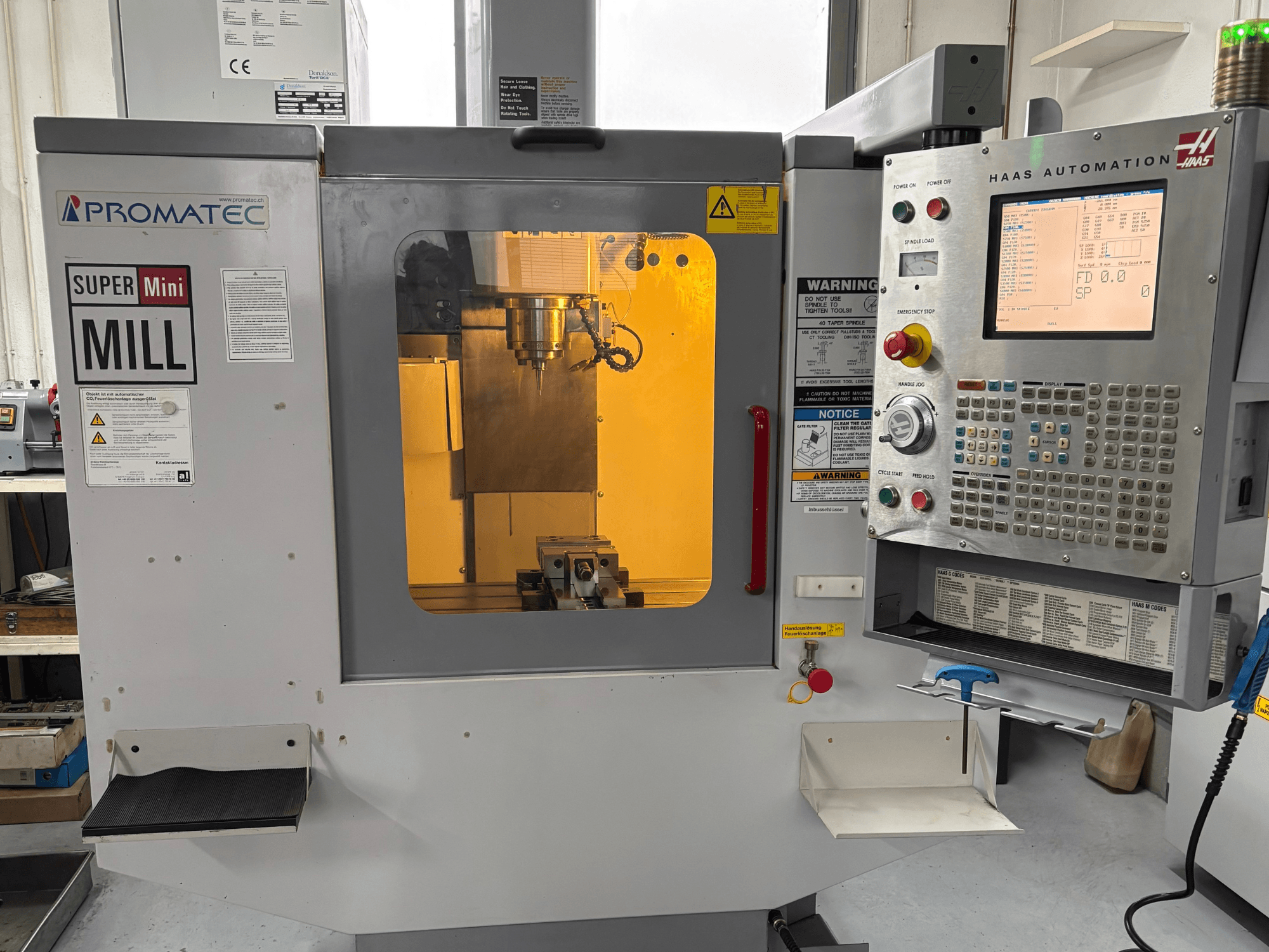Фрезерный станок Promatec Super Mini Mill спереди, оснащенный панелью управления Haas Automation и видимым режущим инструментом внутри.