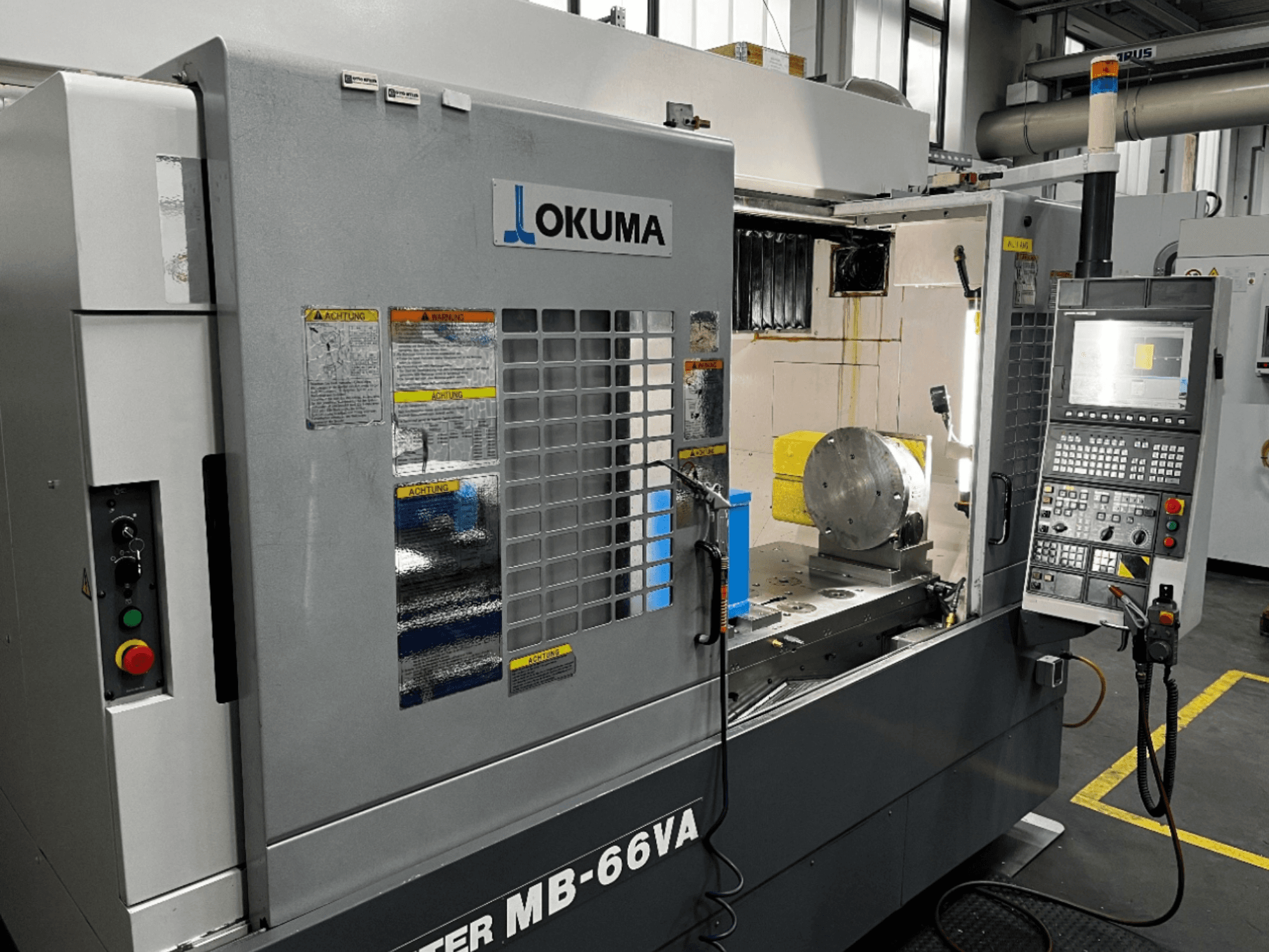 Вид спереди вертикального обрабатывающего центра Okuma MB66-VA с панелью управления, заготовкой и внутренней частью станка с наклейками безопасности.