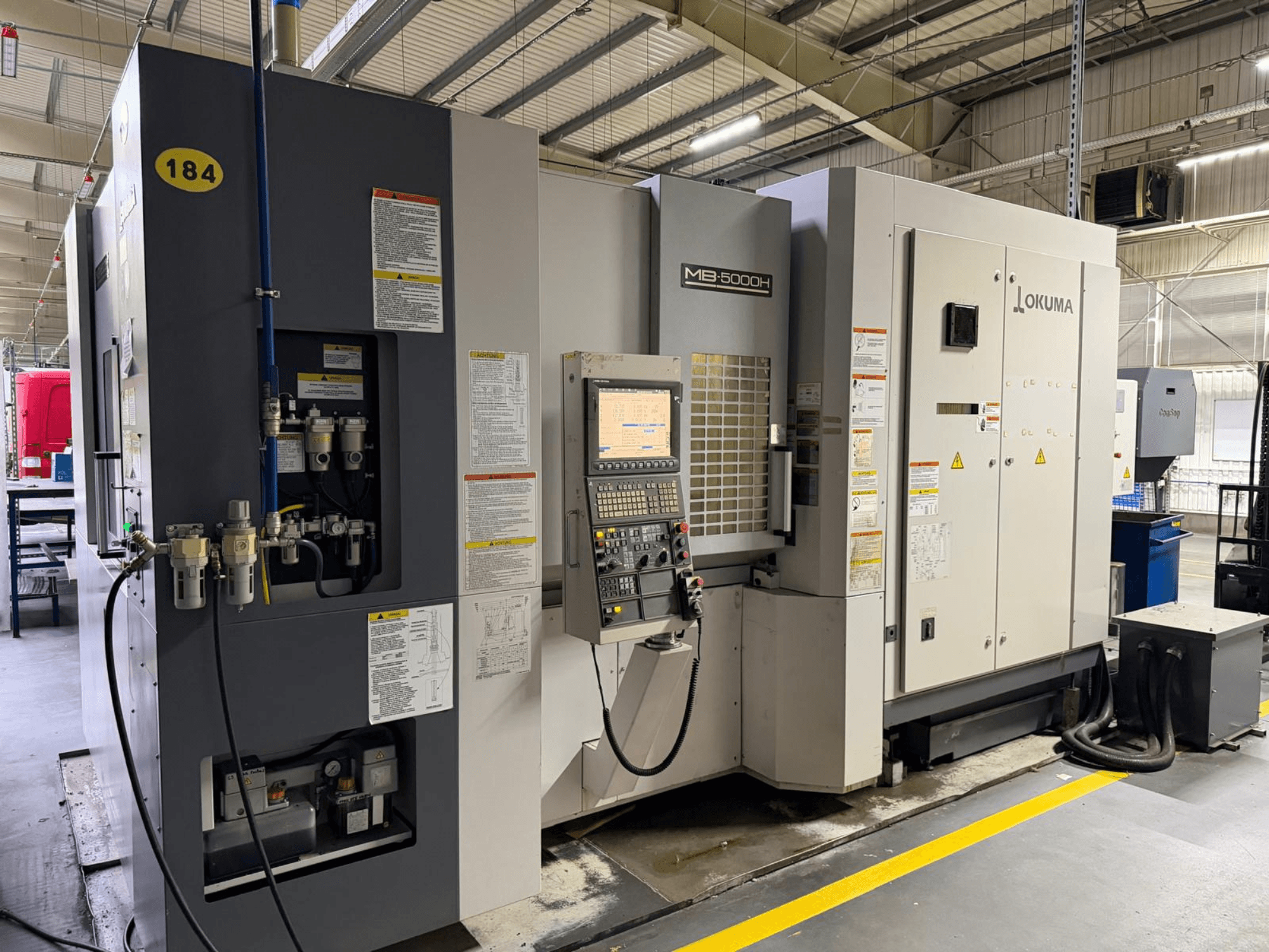 Вид спереди горизонтального обрабатывающего центра Okuma MB 5000H, элементы управления, маркировка и гидравлические соединения.