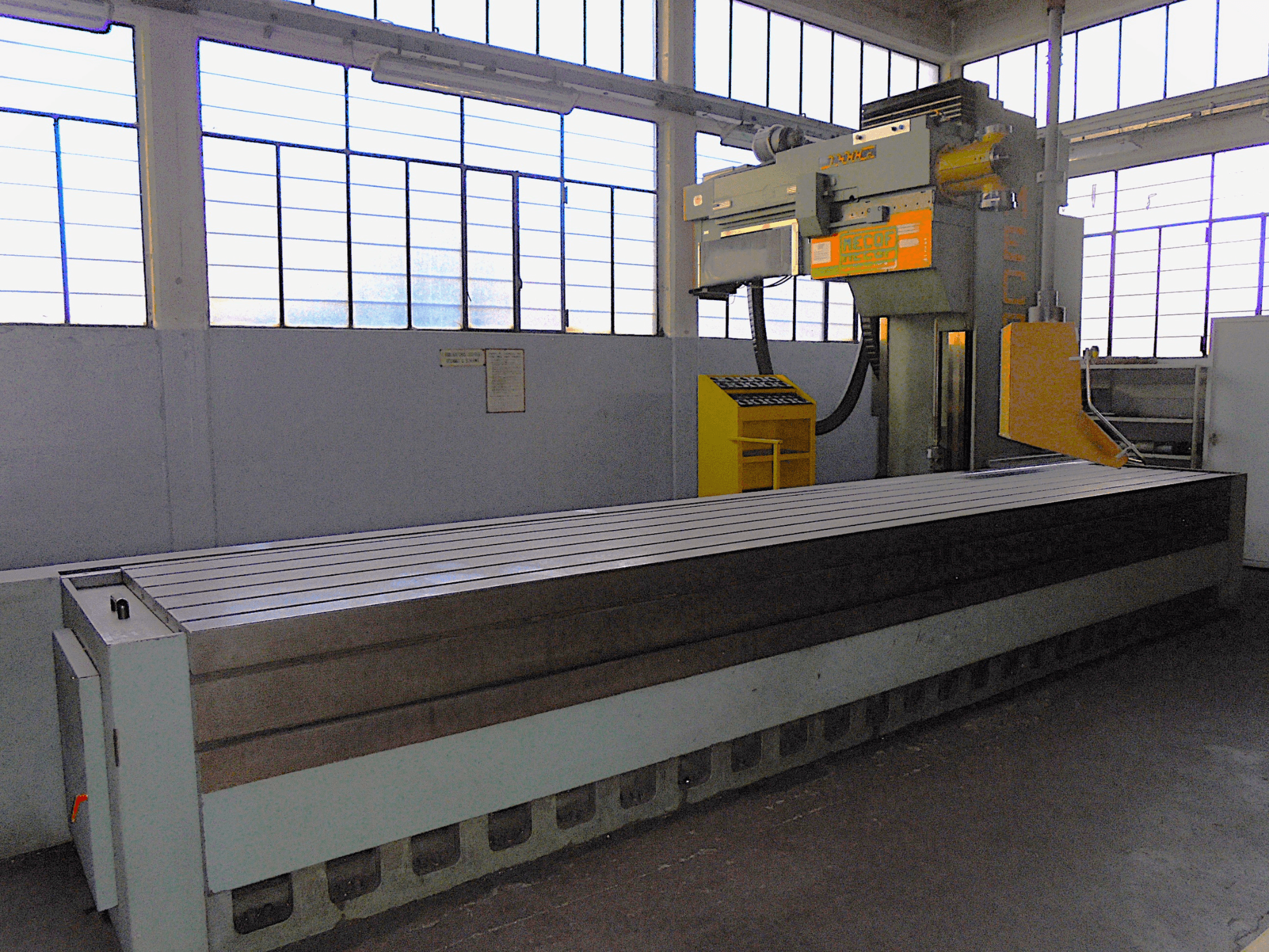 Неизвестная марка, вертикальный фрезерный станок на заводе, вид сбоку, имеет большой стол с рифленой поверхностью и панель управления.