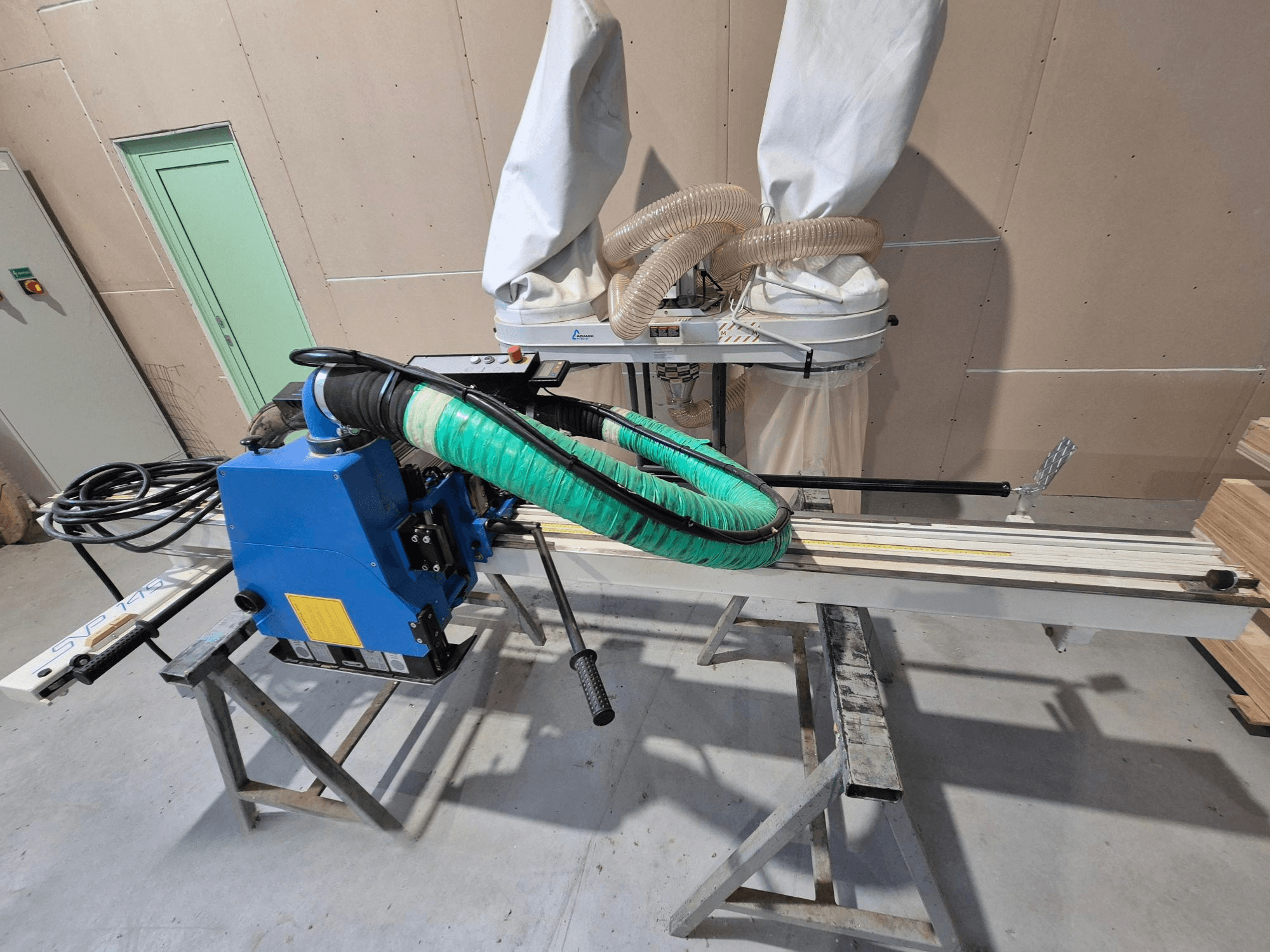 Синяя горизонтальная пила SVP-175 с зеленым пылезащитным шлангом и опорой, вид сбоку в мастерской.