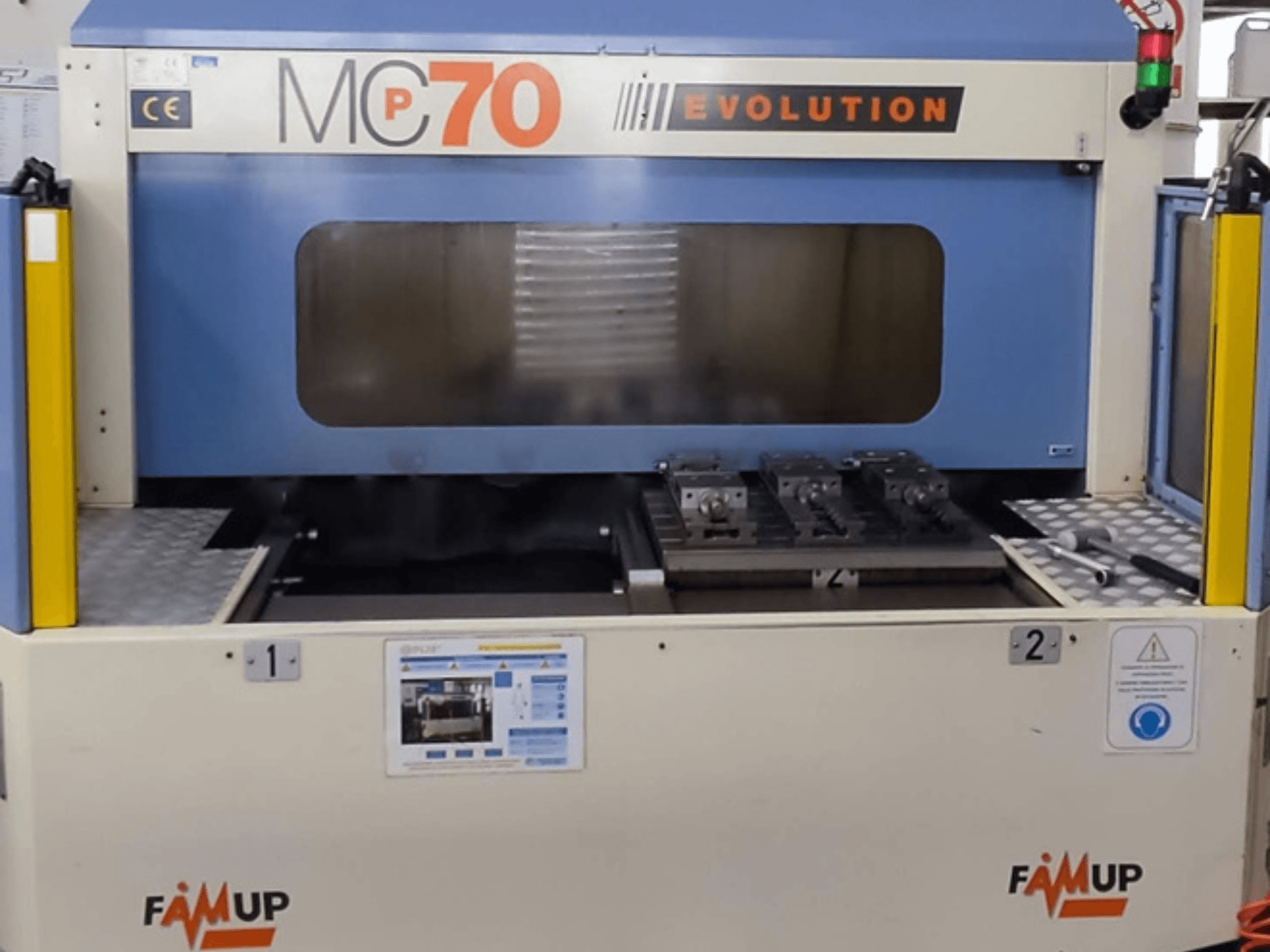 Вертикальный обрабатывающий центр FAMUP MCP70 E, вид спереди, показывающий панель управления и рабочую зону с приспособлениями и средствами безопасности.