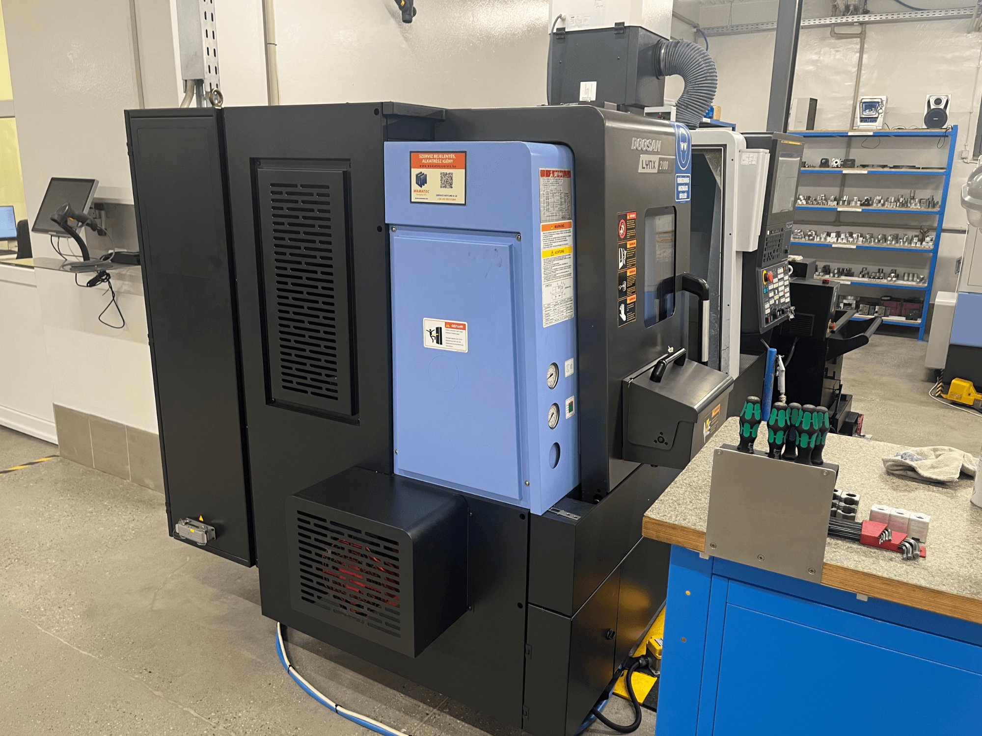 Токарный станок Doosan Lynx 2100, вид сбоку, демонстрирует панель управления, систему вентиляции и инструменты на верстаке.