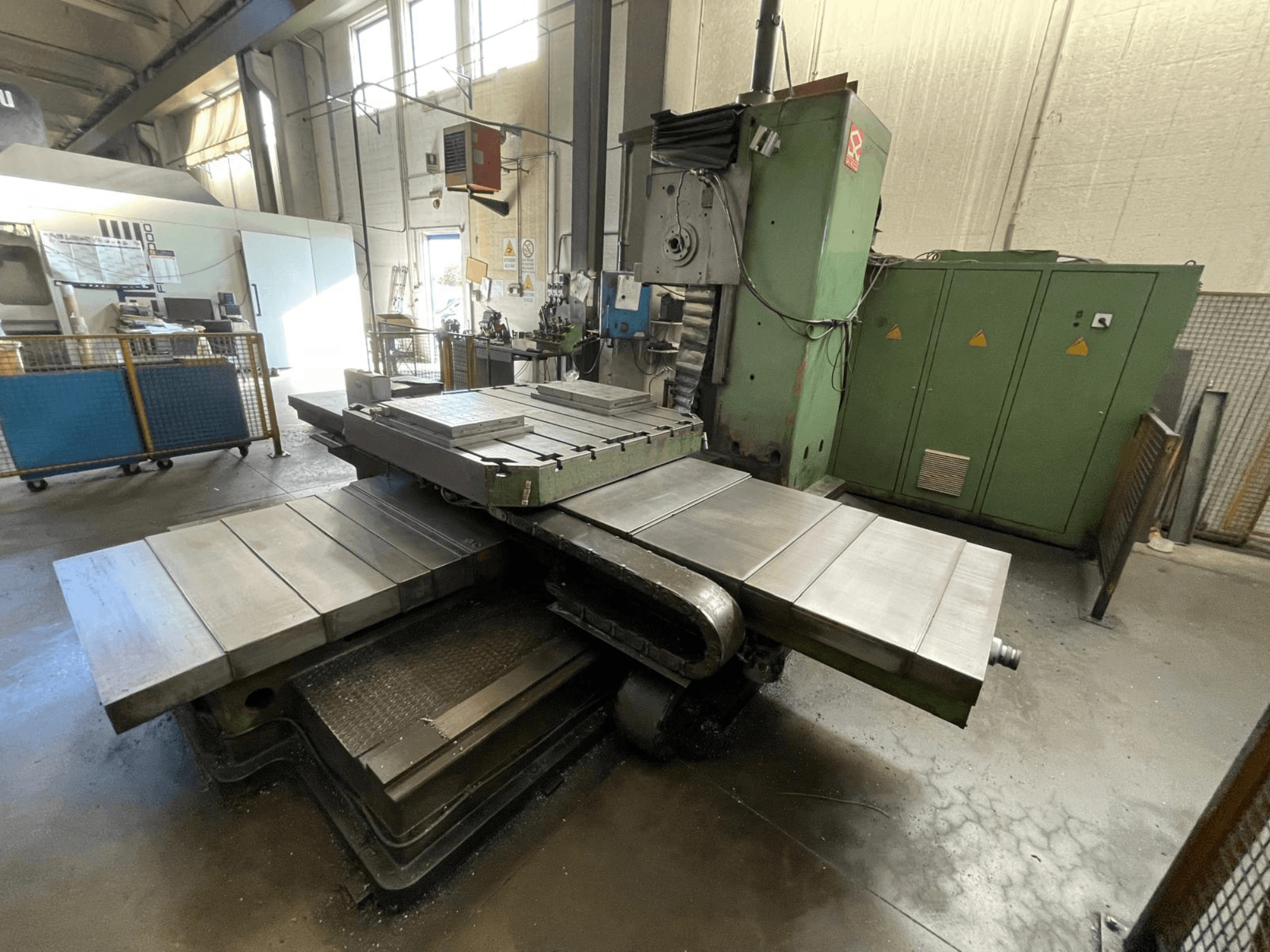 Большой промышленный фрезерный станок с зеленым корпусом, вид сбоку, с несколькими плоскими поверхностями и шкафами для хранения на заднем плане.