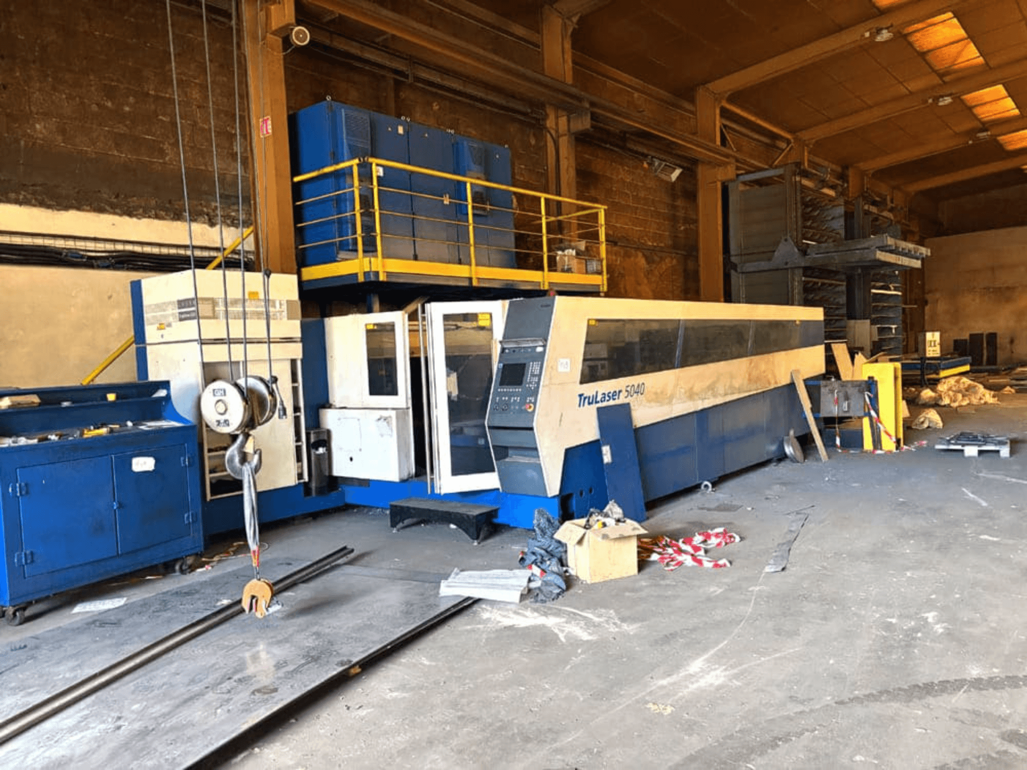 Станок для лазерной резки CO2 производства компании TRUMPF, модель TRULASER 5040, запечатлённый в цехе: вид спереди под углом на фоне заваленного предметами помещения.