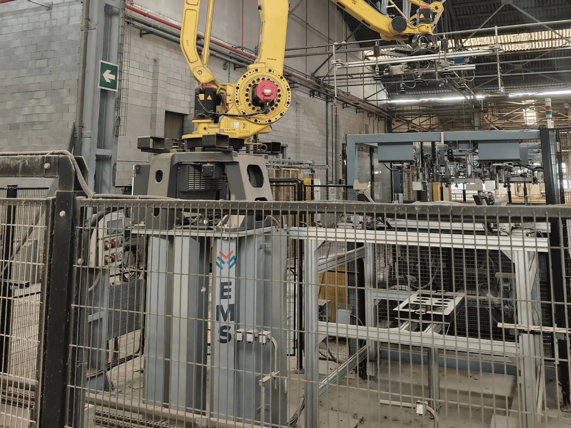 Вид спереди на манипулятор робота FANUC M-410iC/315, установленный над инструментом для монтажа электронных сборок (EMS), на заднем плане — промышленное оборудование и ограждение.