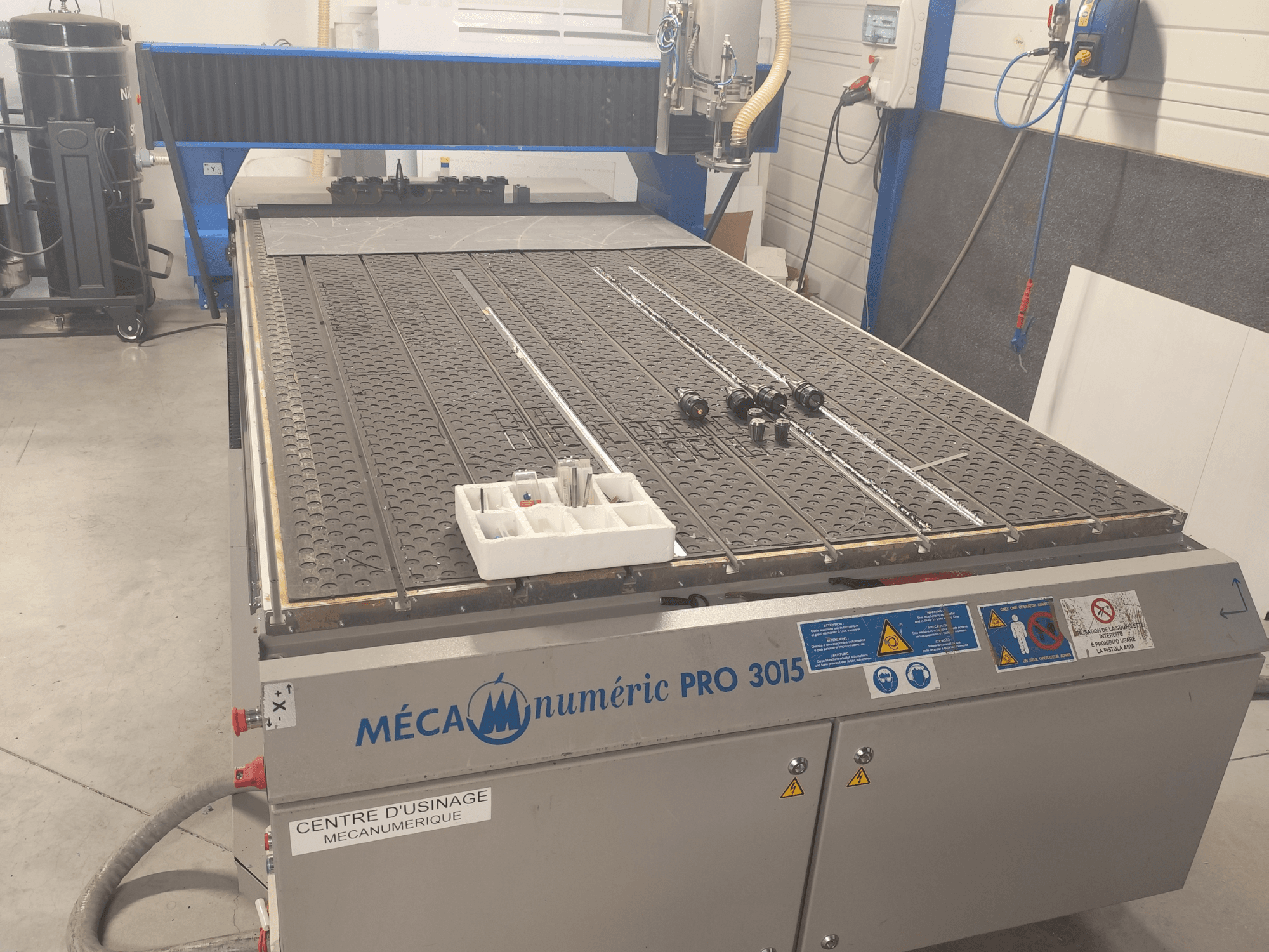 Фрезерный станок с ЧПУ MECANUMERIC Pro 3015, вид спереди, с подробным изображением режущего стола, панели управления и принадлежностей, расположенных на столе.