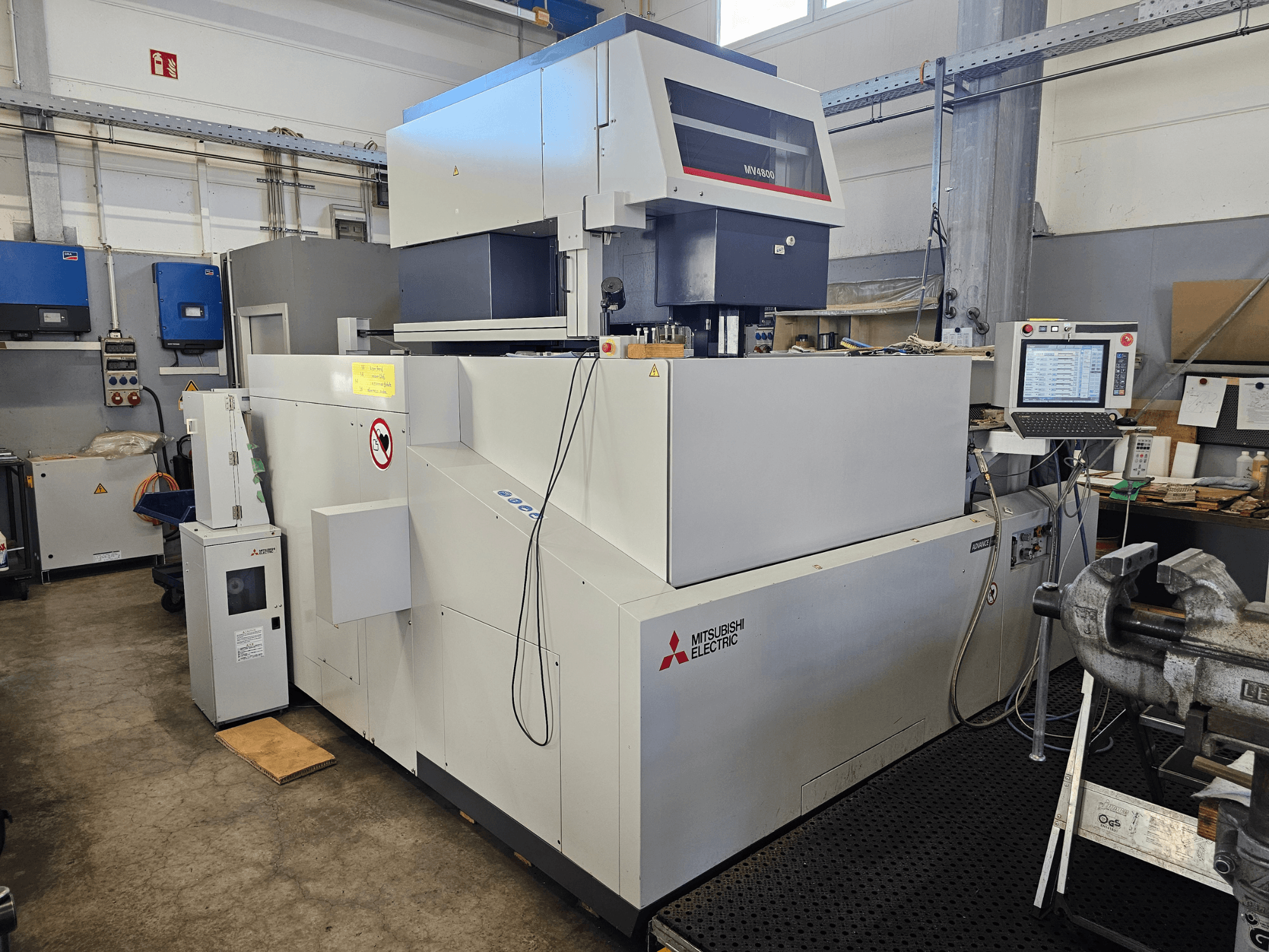 Проволочно-вырезной станок Mitsubishi MV4800 в мастерской, вид сбоку с изображением панели управления, табличек безопасности и окружающего оборудования.