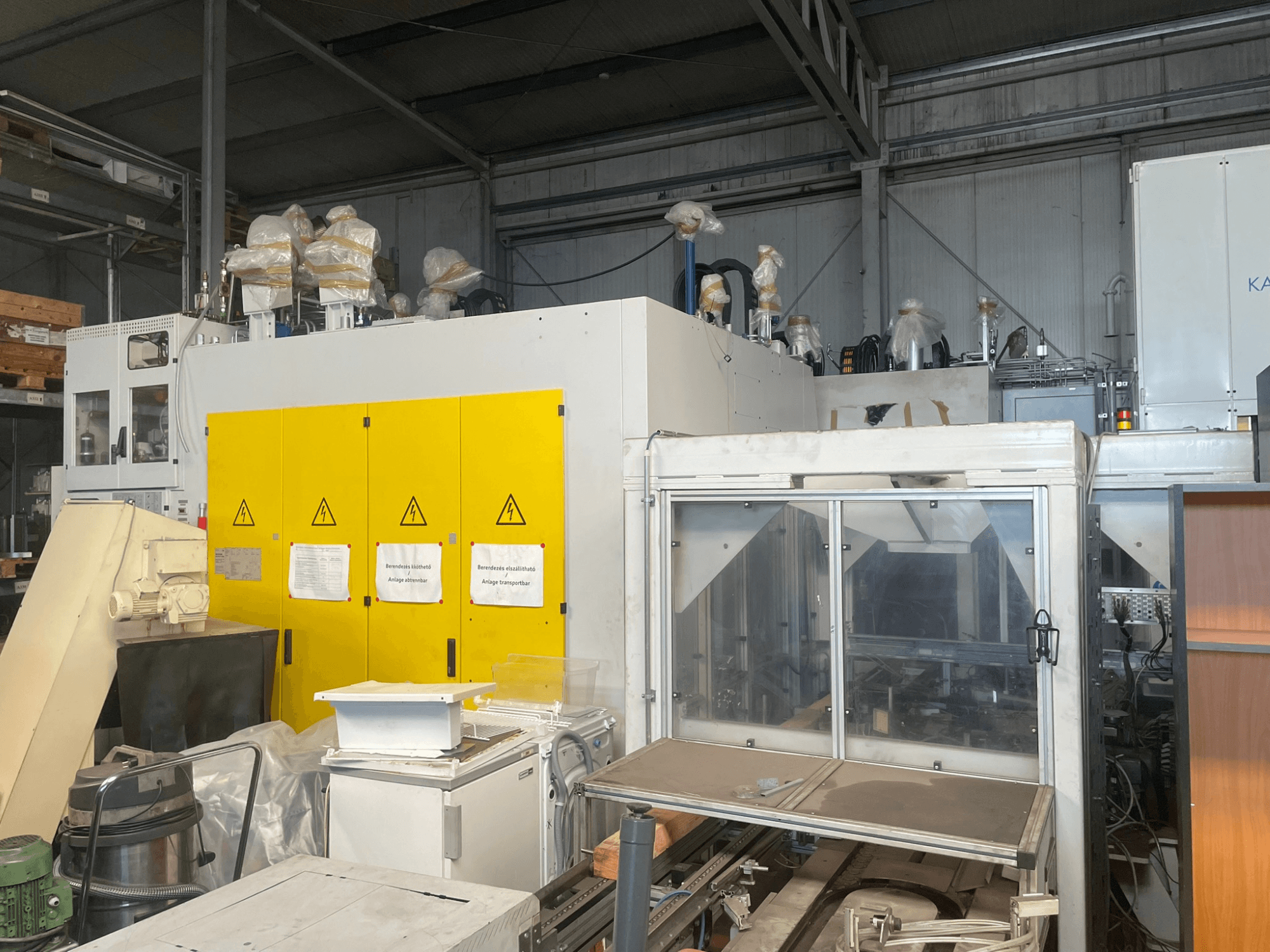 Промышленное оборудование с желтой панелью управления, вид сбоку, с предупреждающими надписями и пластиковыми накладками, на складе.