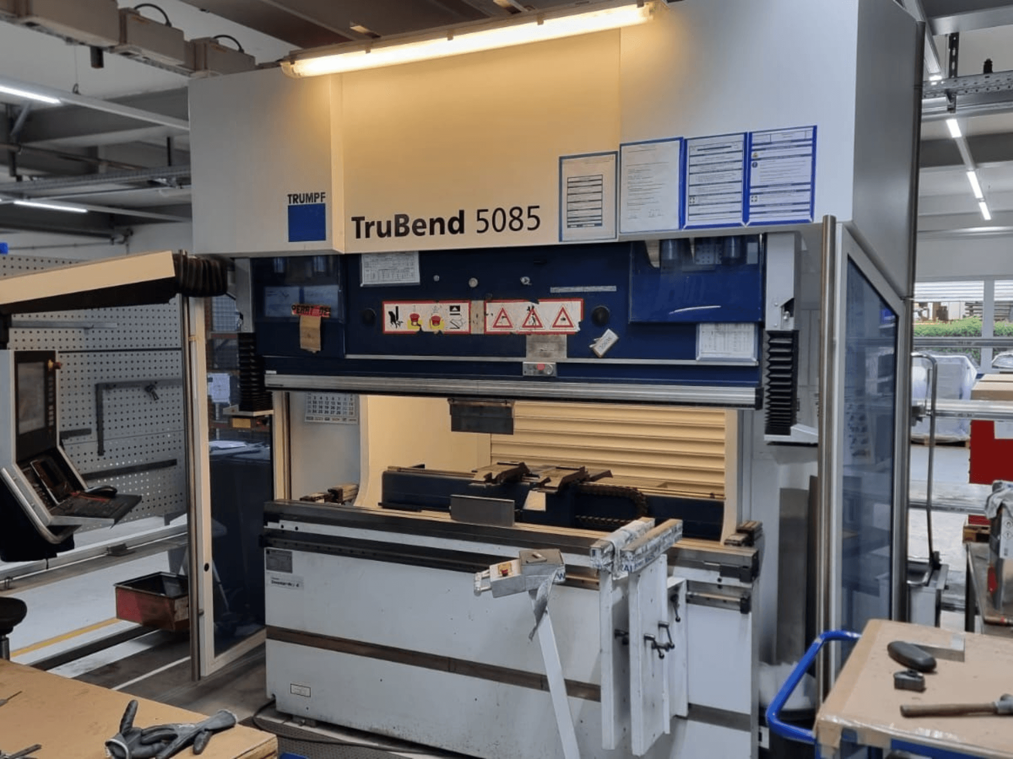 Гибочный станок TRUMPF TruBend 5085, вид спереди, панель управления, рабочая станция и предупреждающие знаки безопасности в мастерской.