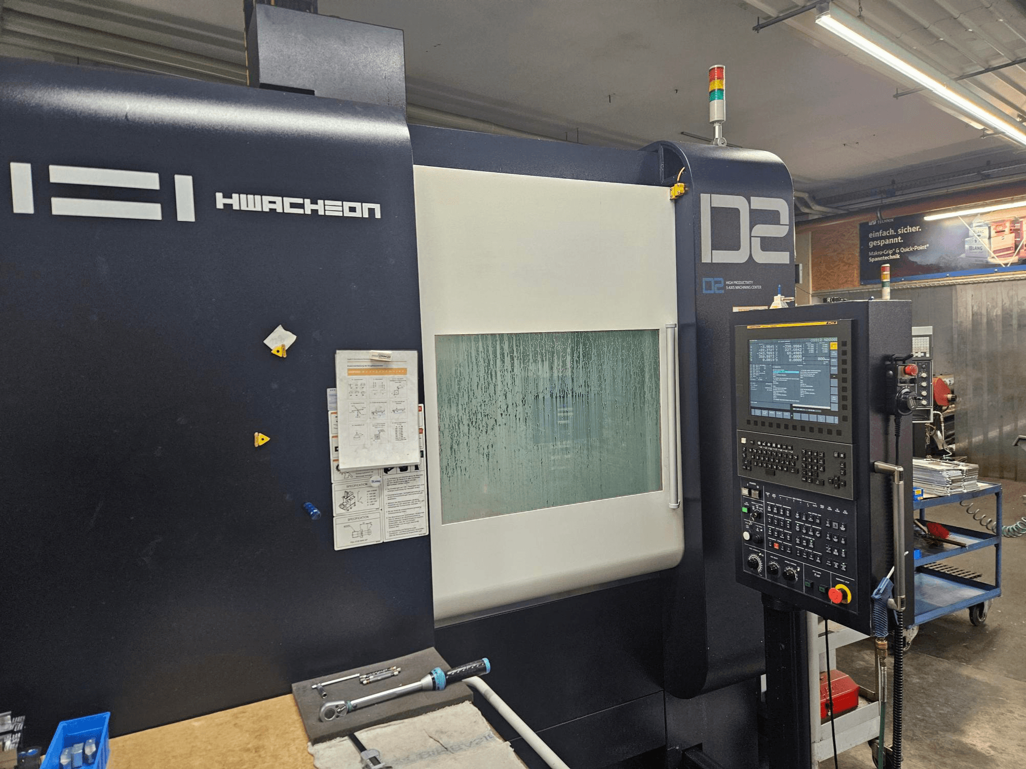 Вертикальный обрабатывающий центр HWACHEON D2-5AX 12k, вид спереди с панелью управления и запотевшим смотровым окном.