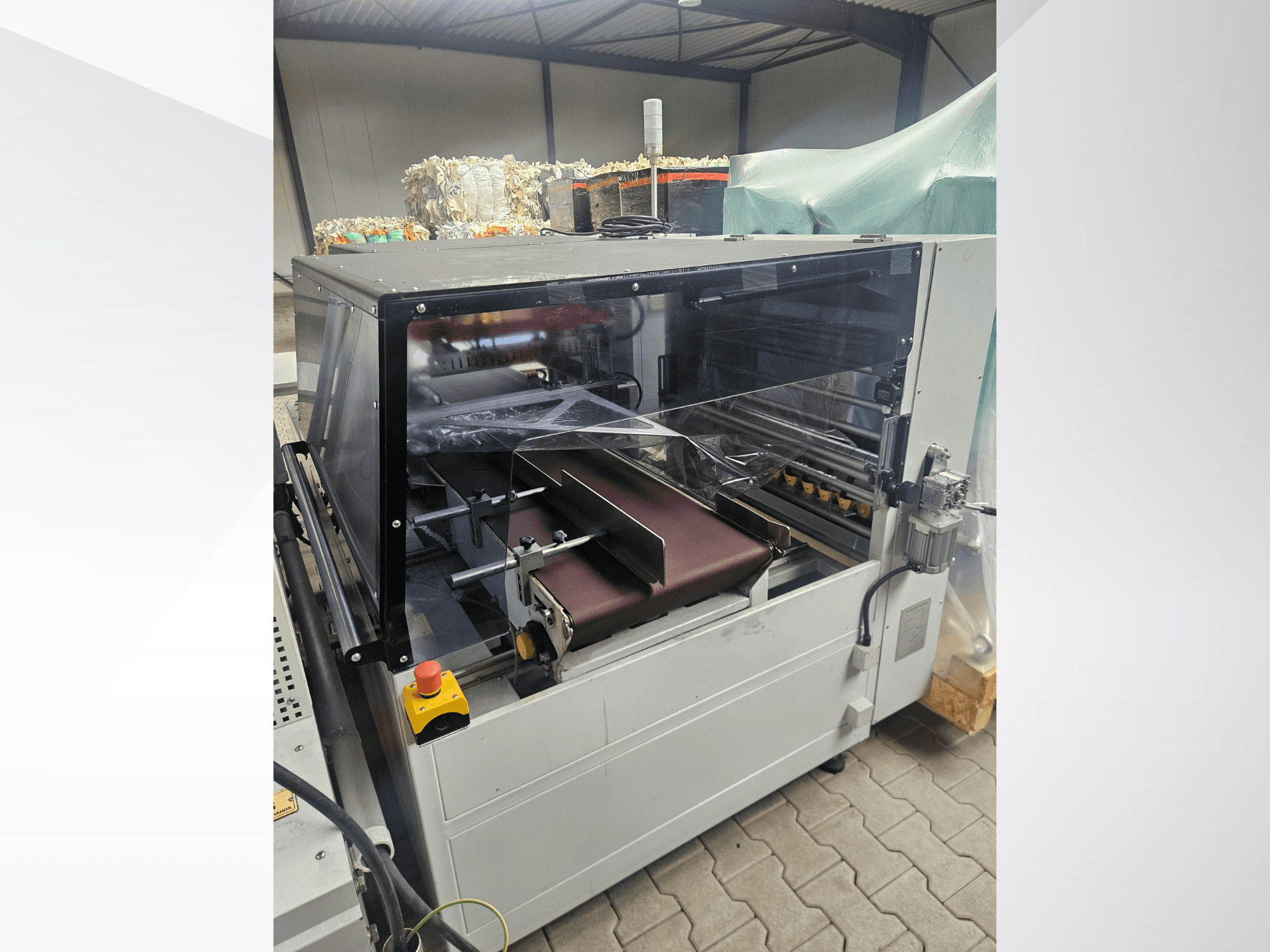 Машина AMTEC Speed Shrink 40 PRO, вид спереди, на котором виден механизм обработки пластика с прозрачной защитной крышкой и конвейерной лентой.
