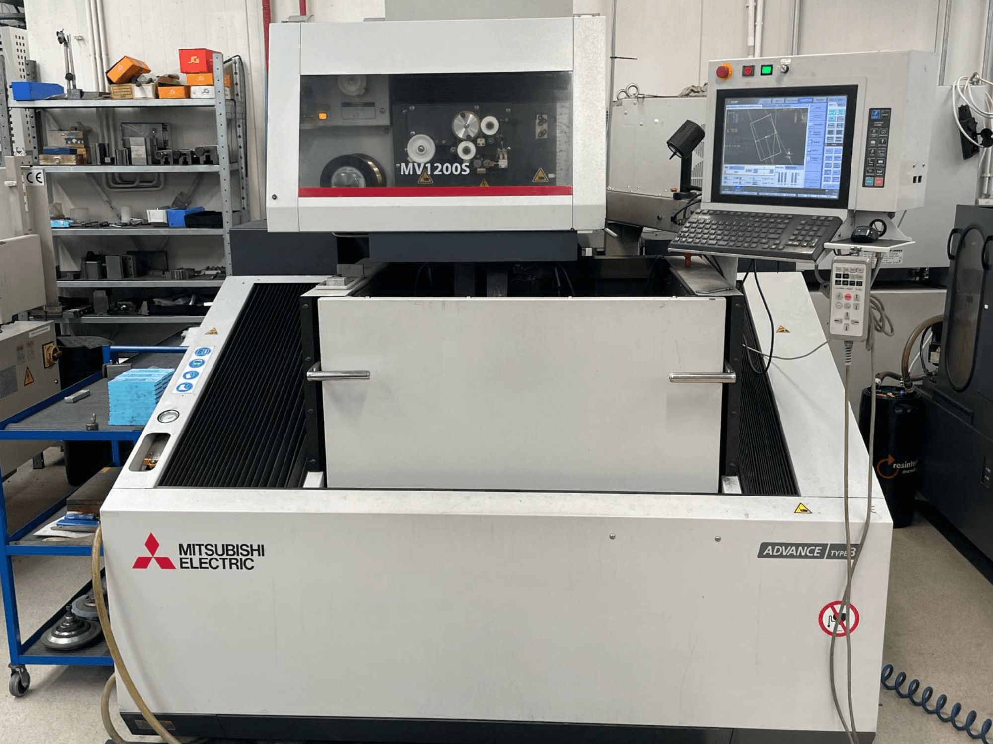 Электроэрозионный станок Mitsubishi MV1200S в виде спереди, на котором видны панель управления и рабочая зона, а также предупреждающие надписи о мерах безопасности.