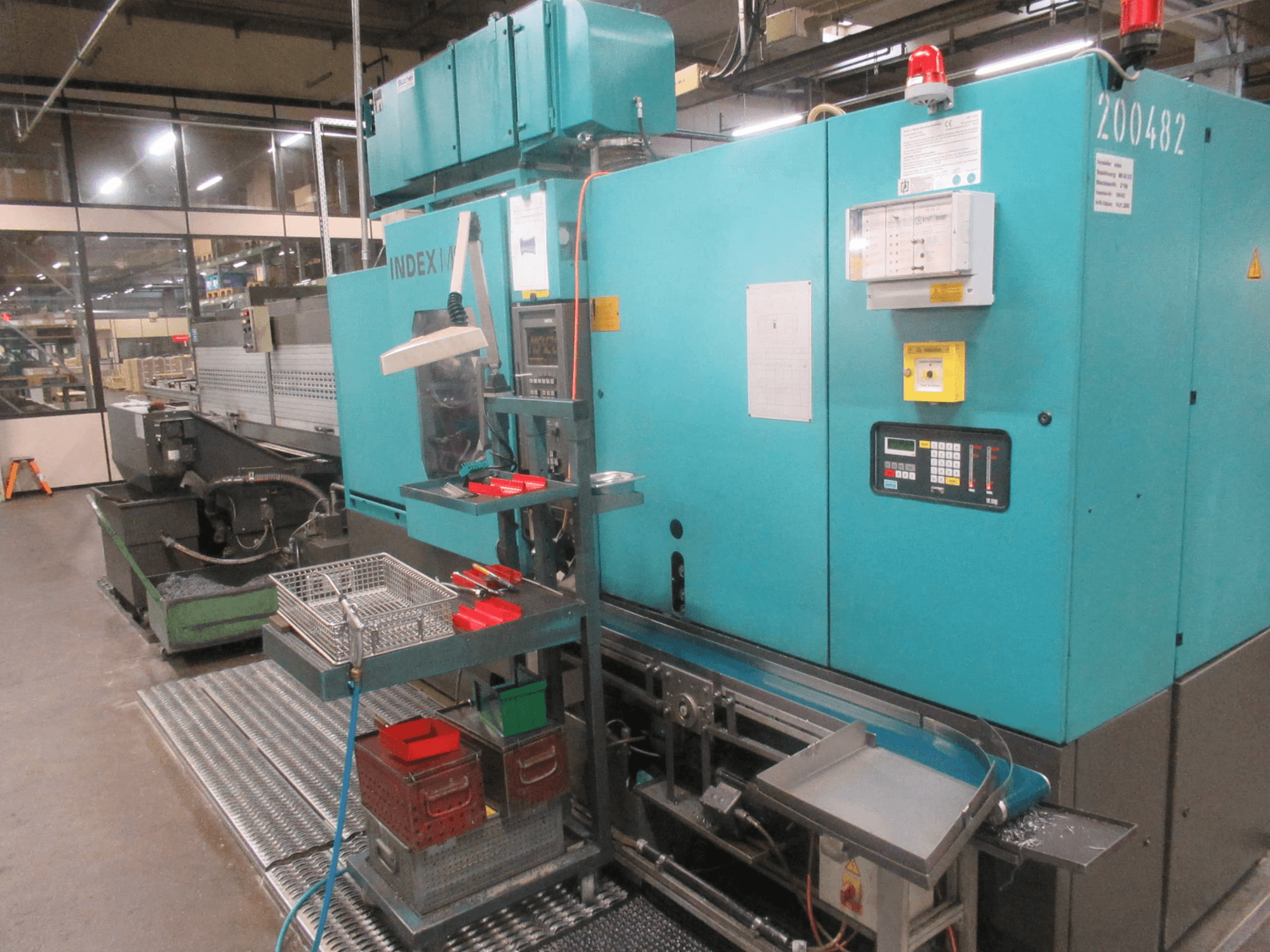 Бирюзовый станок INDEX на заводе, вид сбоку; панель управления, хранилище инструментов и конвейер. Видны бункеры, шланги и сигнализация.