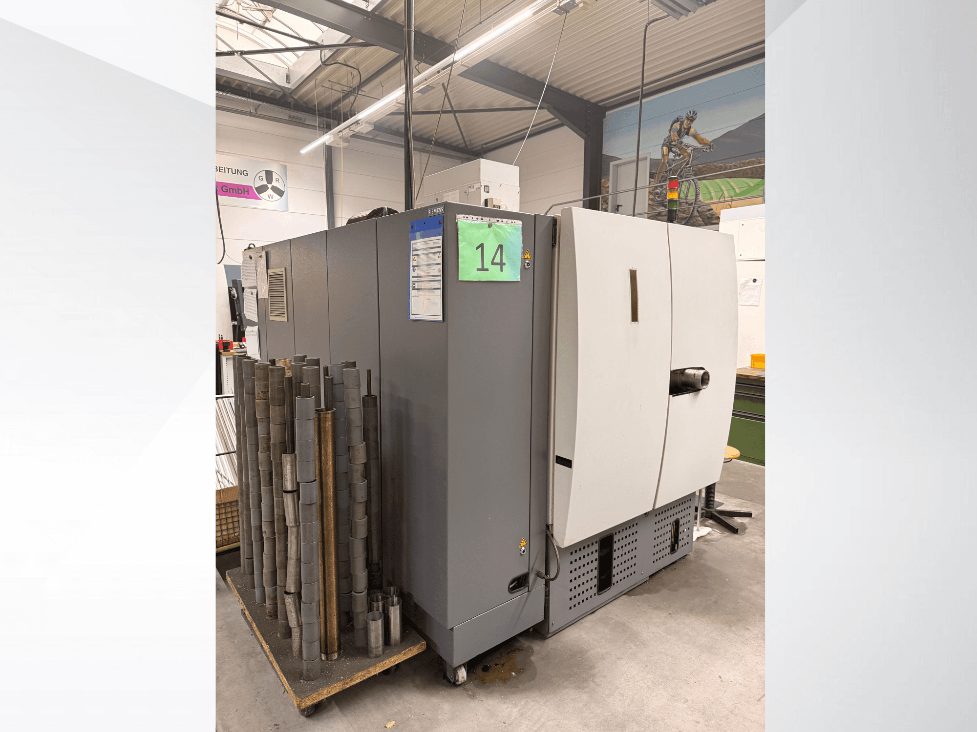 Серый промышленный станок Siemens, вид сбоку, с запасом цилиндрических металлических деталей рядом с ним и видимыми этикетками оборудования.