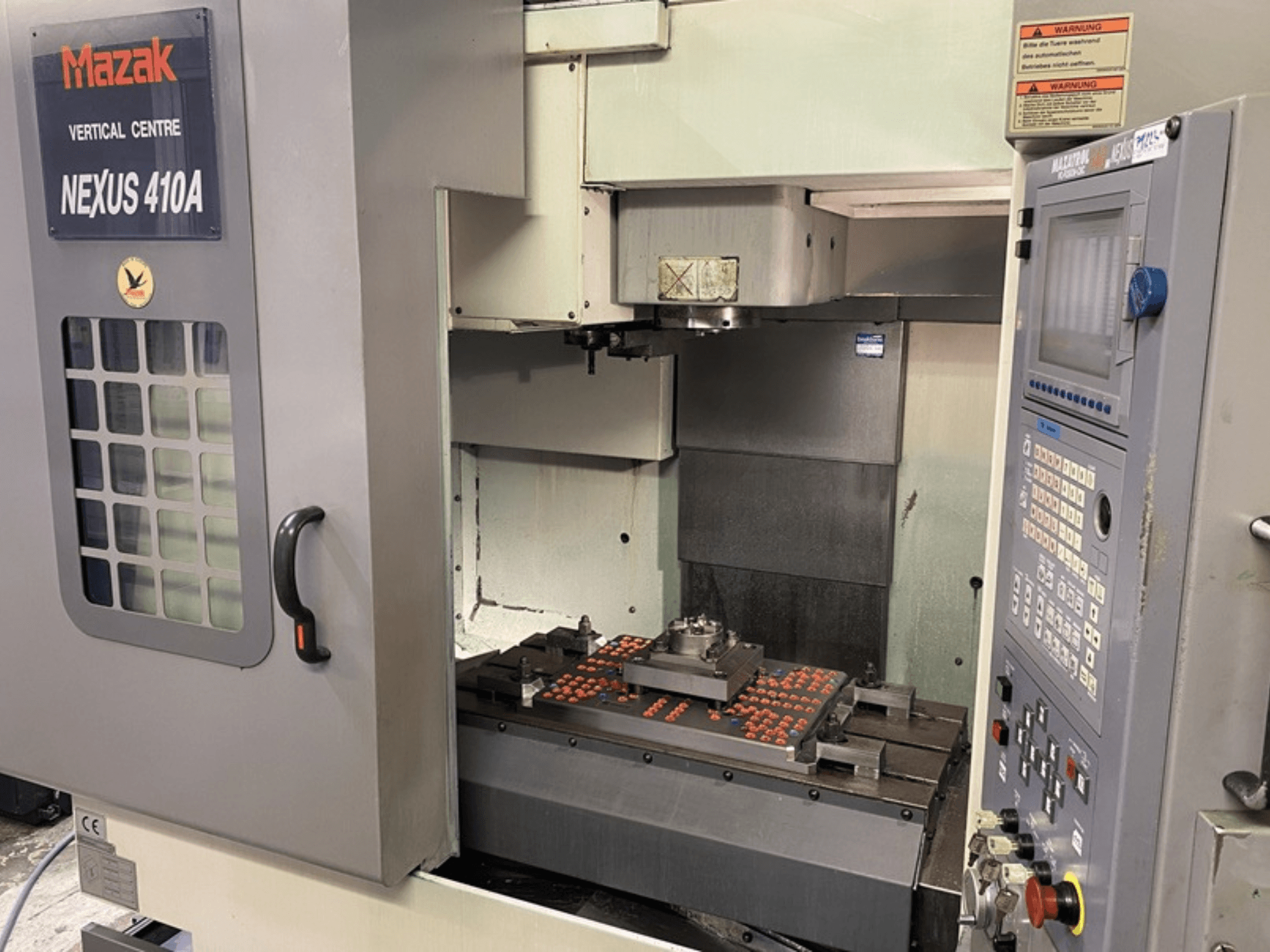 Вертикальный обрабатывающий центр Mazak VCN410, вид спереди, показывающий панель управления и внутреннюю часть станка с установленной заготовкой.