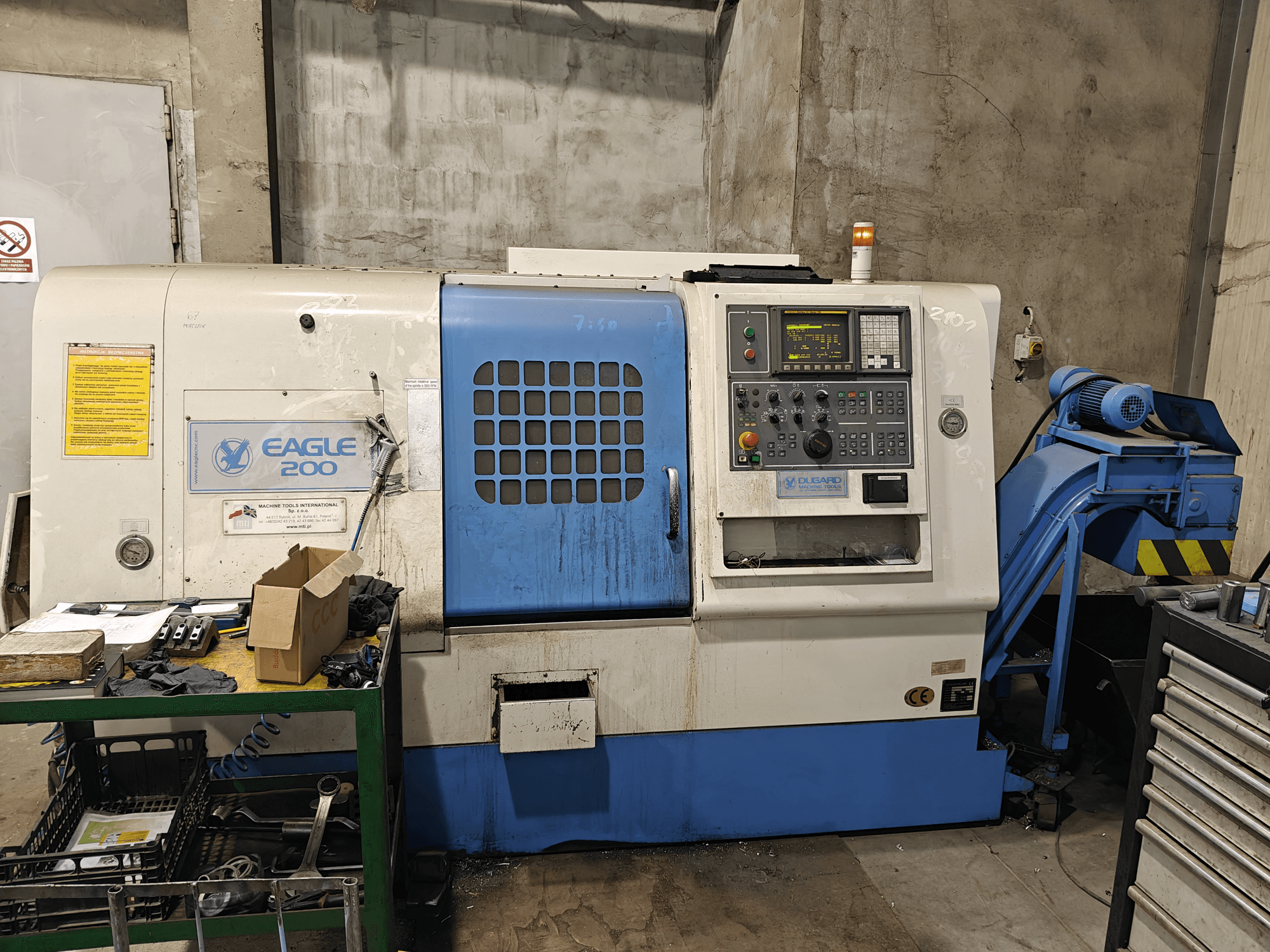 Станок с ЧПУ Eagle 200, вид спереди с панелью управления, различные инструменты на зеленой тележке. Видимый износ и рабочие индикаторы.