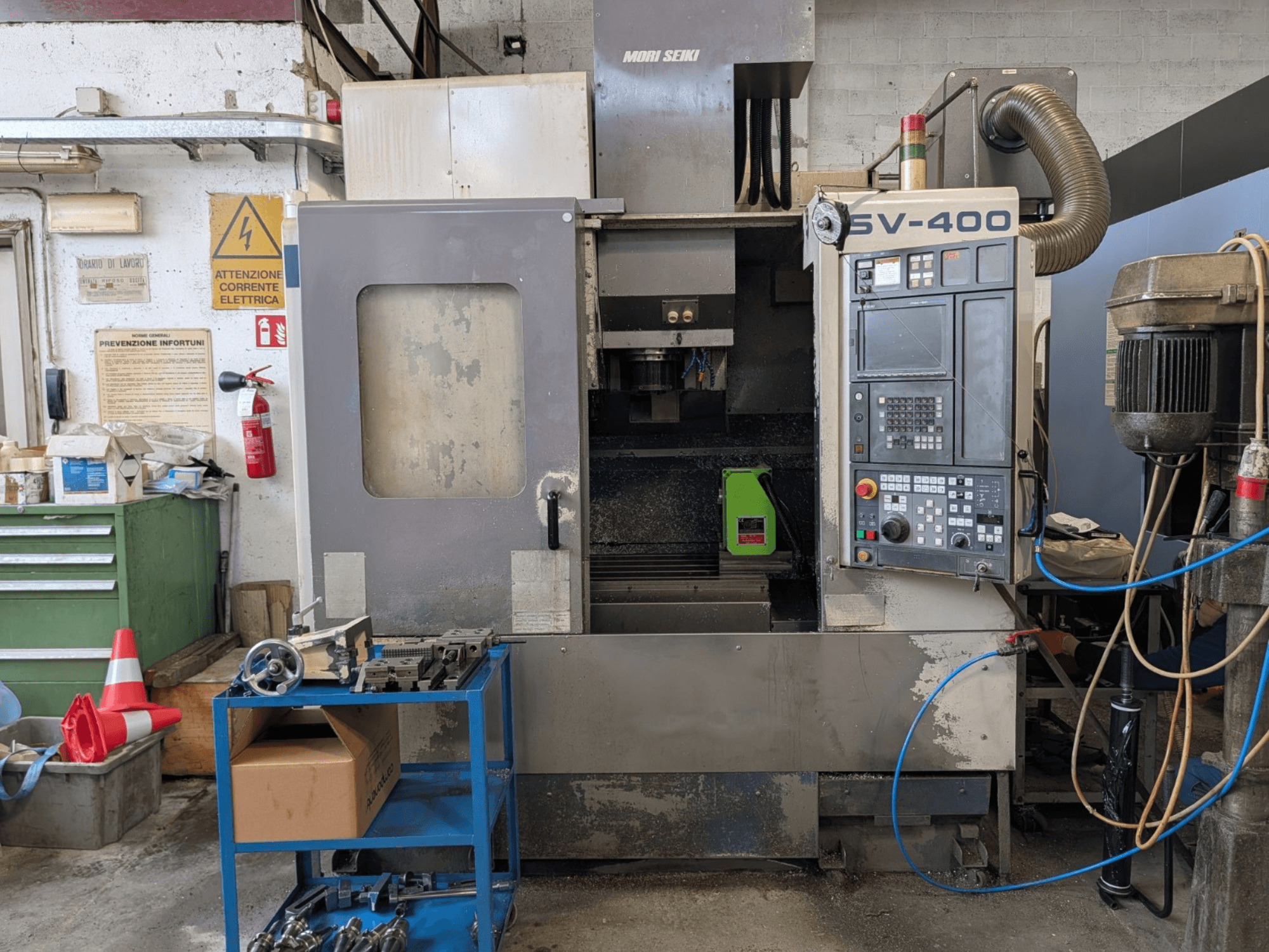 Вертикальный обрабатывающий центр MORI SEIKI SV-400 в мастерской, вид сбоку, видна панель управления и станки рядом.