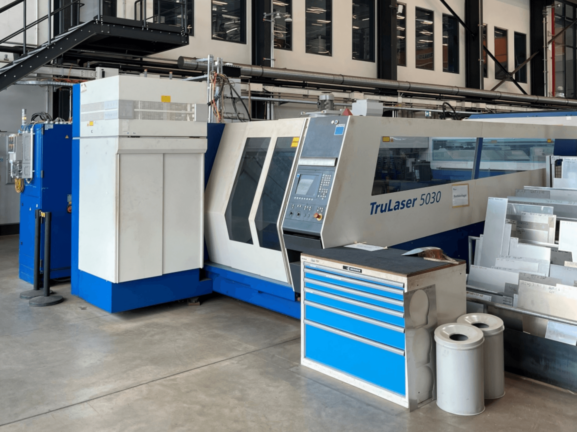 Лазерный резак TRUMPF TruLaser 5030 с CO₂-лазером, вид спереди, с панелью управления и расположенным рядом рабочим столом с металлическими листами.