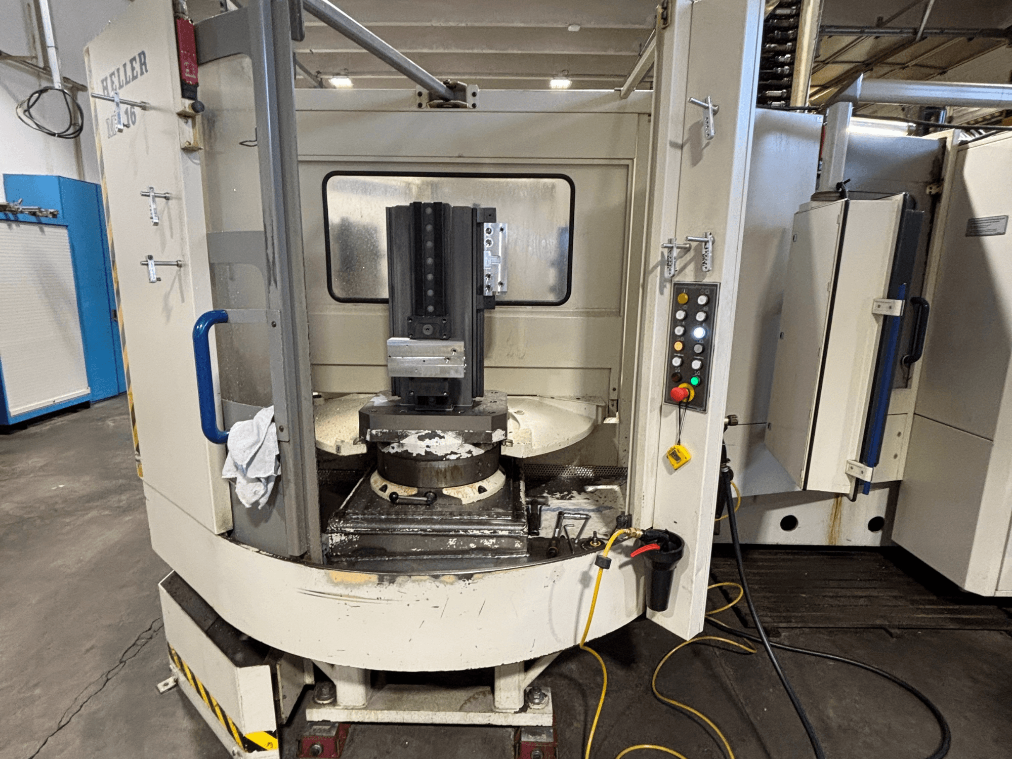 Обрабатывающий центр Heller, модель вертикальная, вид спереди, показывающий шпиндель, панель управления и рабочий стол с установленным оборудованием.