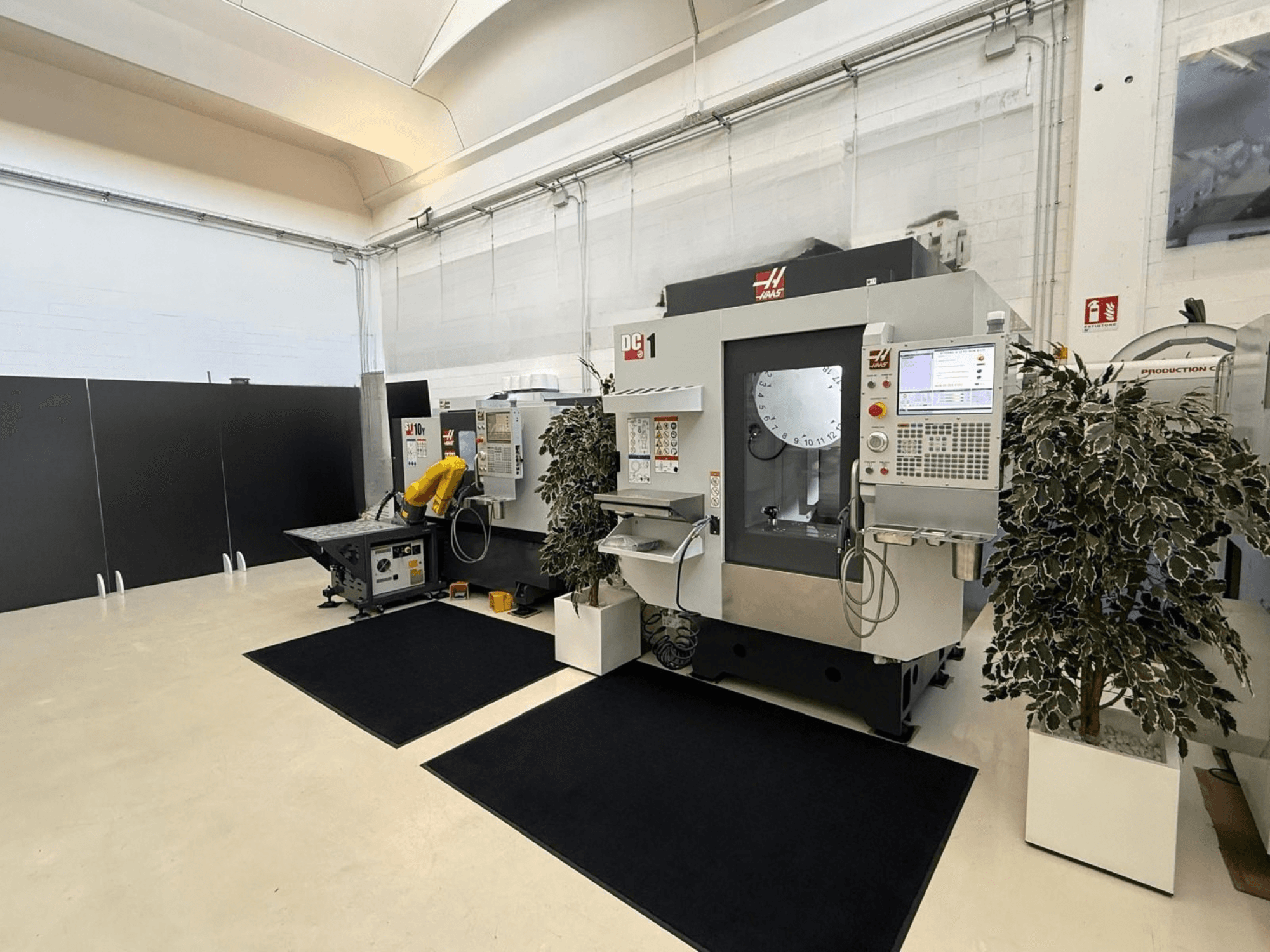Вид спереди на вертикальный обрабатывающий центр HAAS DC-1 в чистом производственном помещении с сенсорной панелью управления и окружающим оборудованием.