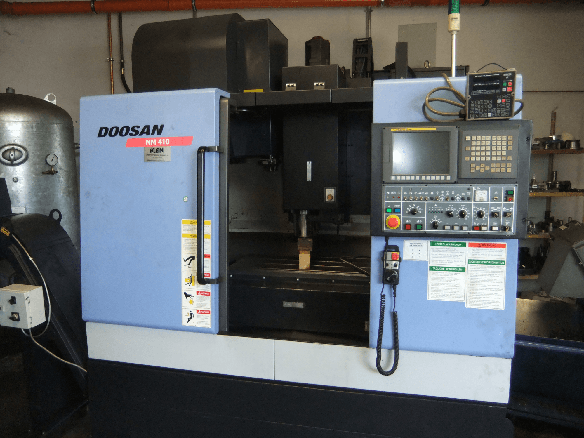 Вертикальный обрабатывающий центр DOOSAN NM 410, вид спереди, на котором видны панель управления, зона шпинделя и предупреждающие надписи.