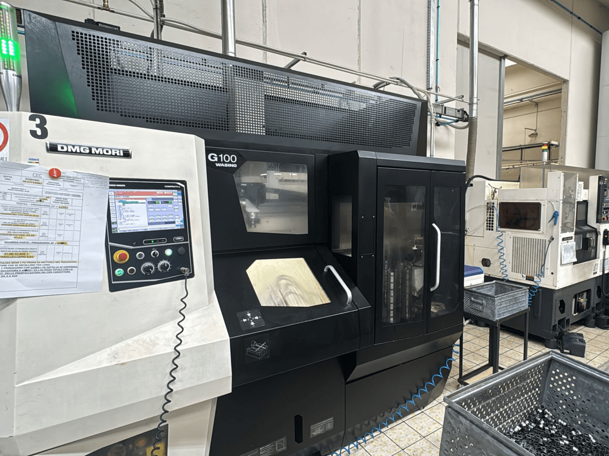 Станок DMG MORI G100 на заводе, вид спереди, с цифровой панелью управления и расположенными рядом контейнерами для хранения инструментов.