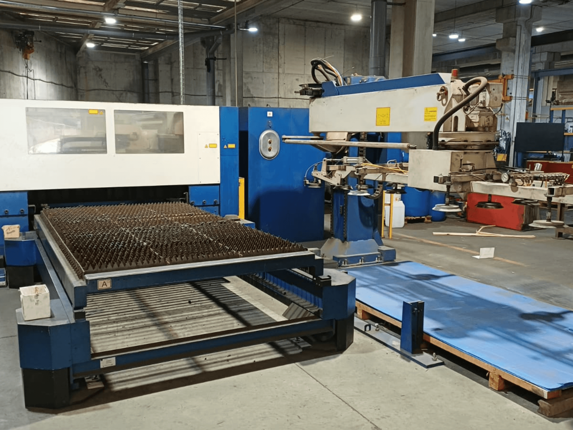 Лазерный резак TRUMPF TRUMATIC L 3030 с CO₂-лазером, вид сбоку, на котором виден режущий стол с шипованной поверхностью и манипулятор.