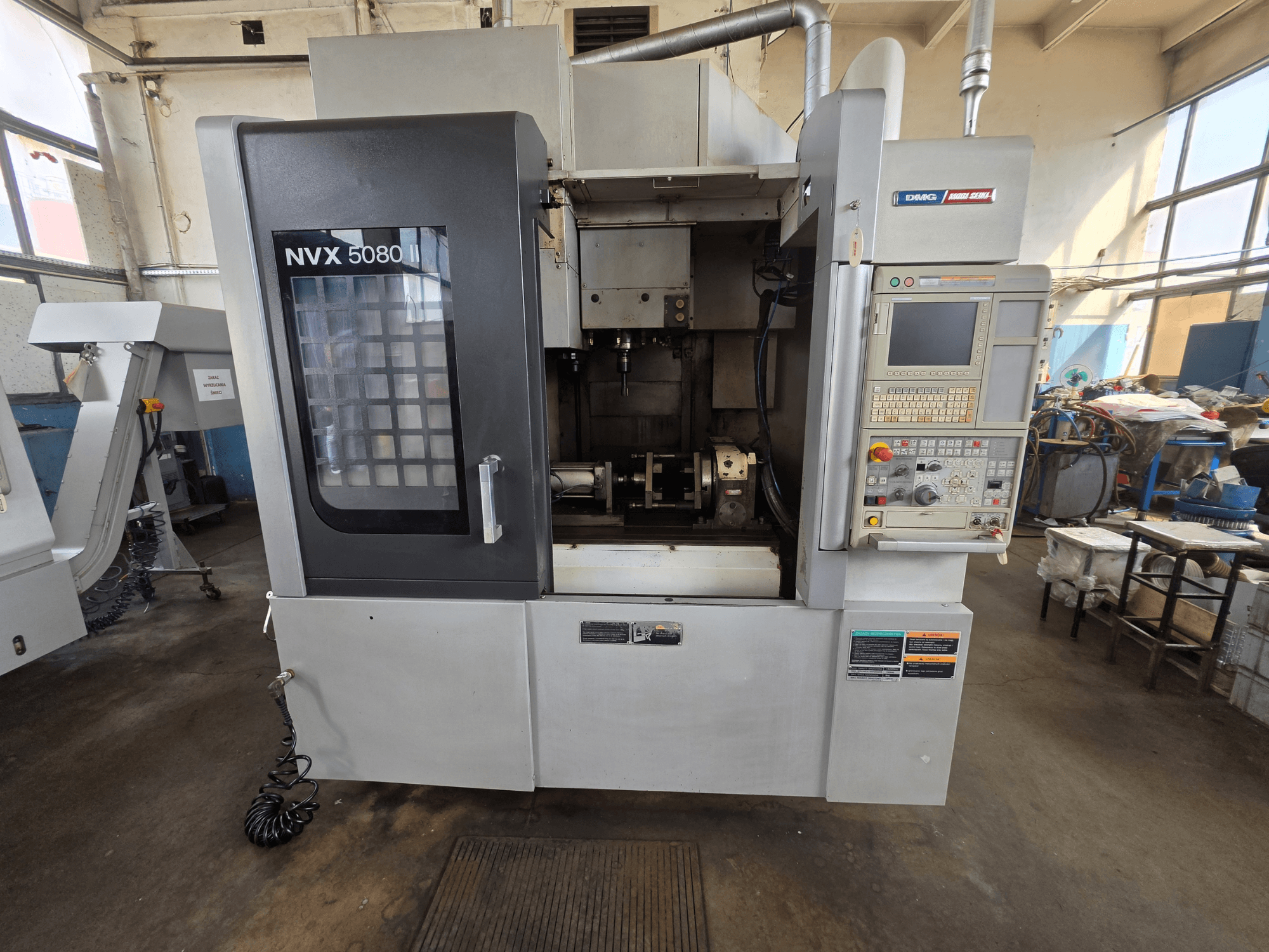 Вид спереди на вертикальный обрабатывающий центр DMG Mori Seiki NVX 5080 II/40 с панелью управления и прозрачным окном, через которое видна зона обработки.