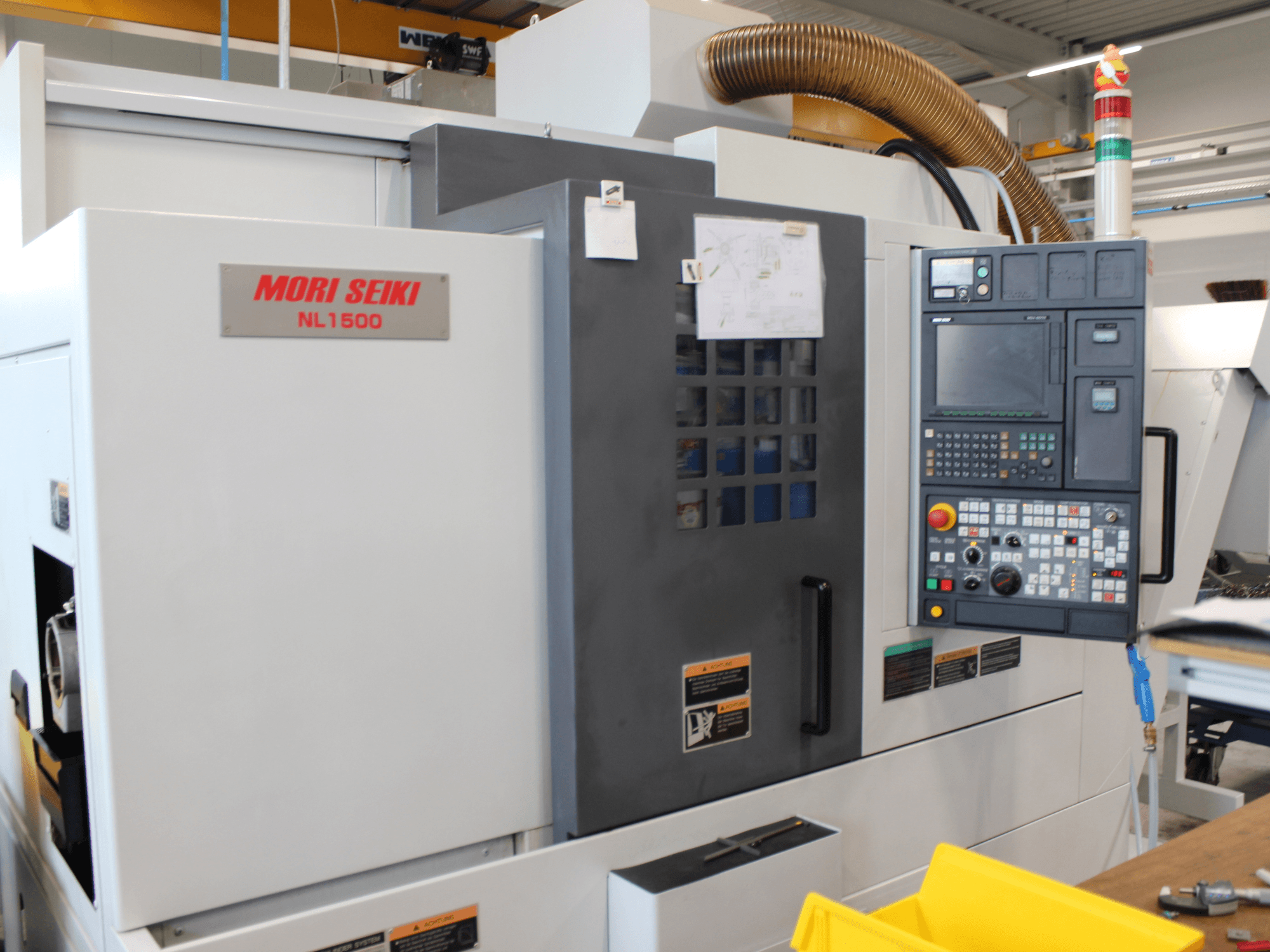 Токарно-фрезерный центр MORI SEIKI NL 1500 SY в боковом виде с панелью управления, знаками техники безопасности и системой охлаждения.