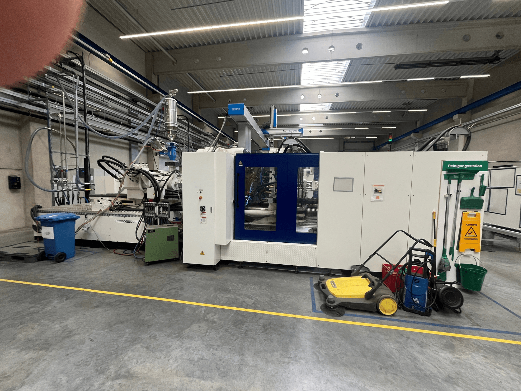 Промышленный станок на заводе, вид спереди, с синими и белыми панелями, шлангами и чистящим оборудованием рядом.