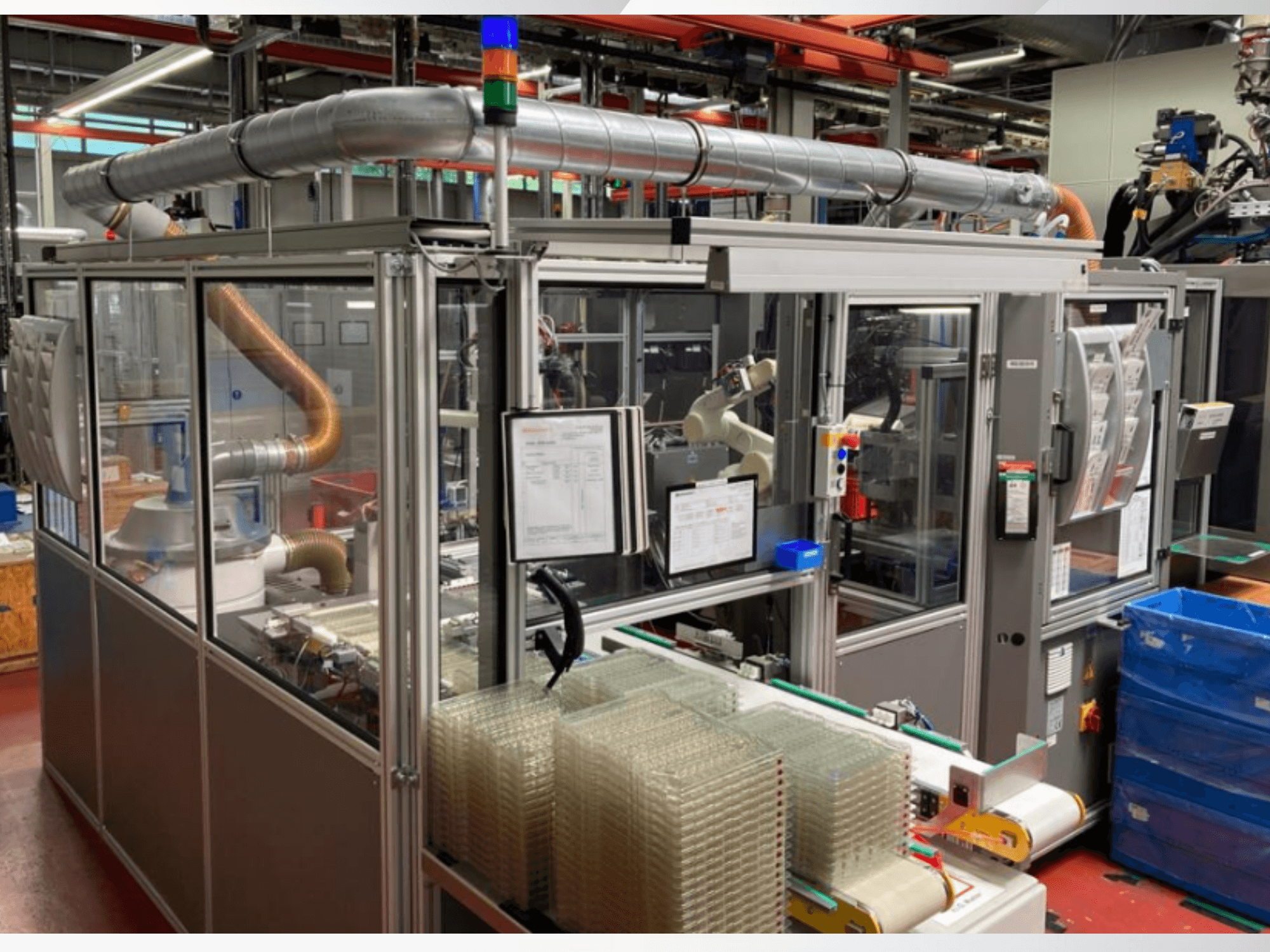 Вид спереди на автоматизированную машину с металлическими трубами, панелью управления и штабелями прозрачных пластиковых контейнеров на заводской площадке.