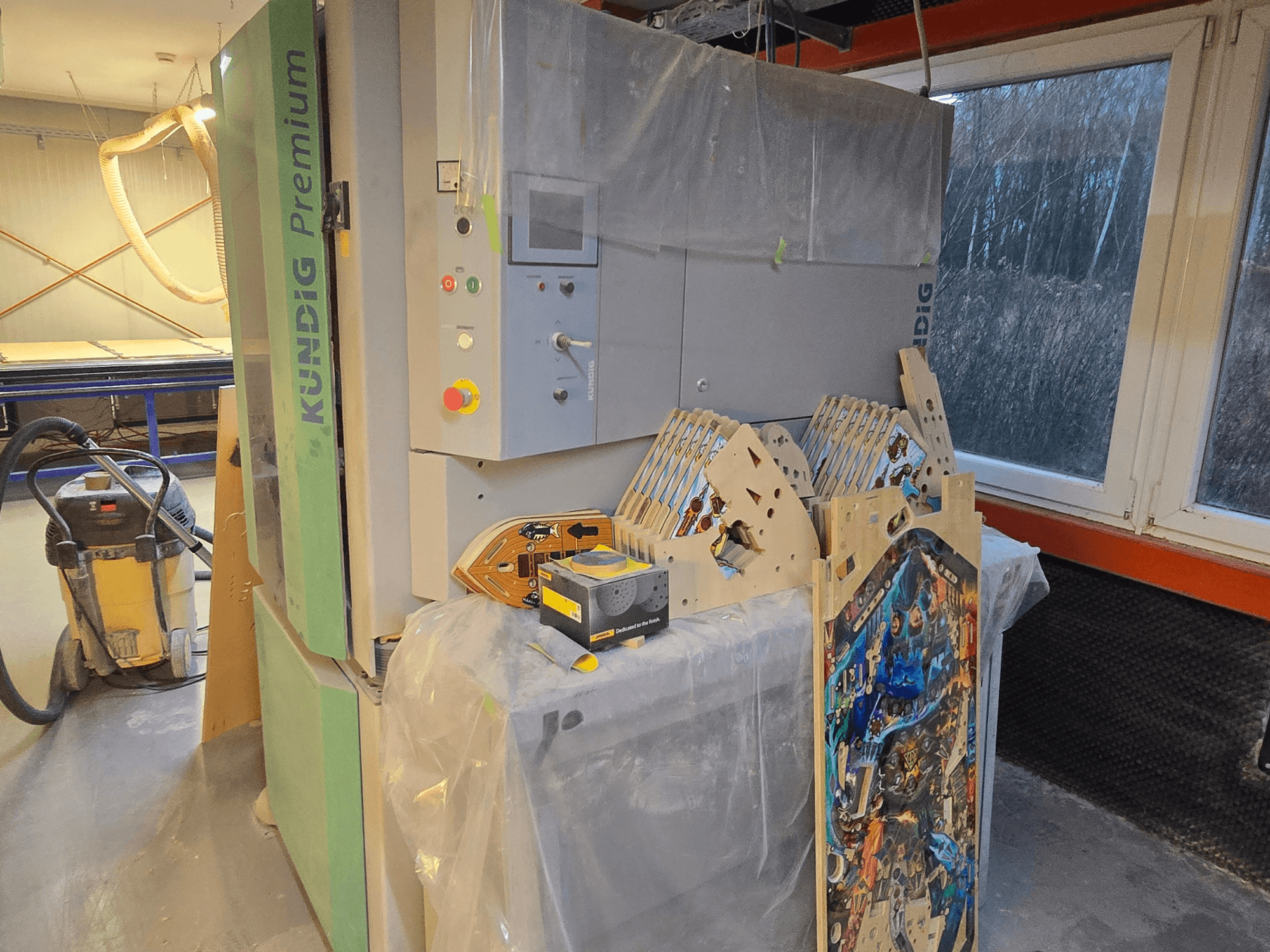 Ленточная шлифовальная машина Kundig Premium-1 1100-C в мастерской, панель управления и деревянные детали, вид сбоку, пылесос на заднем плане.