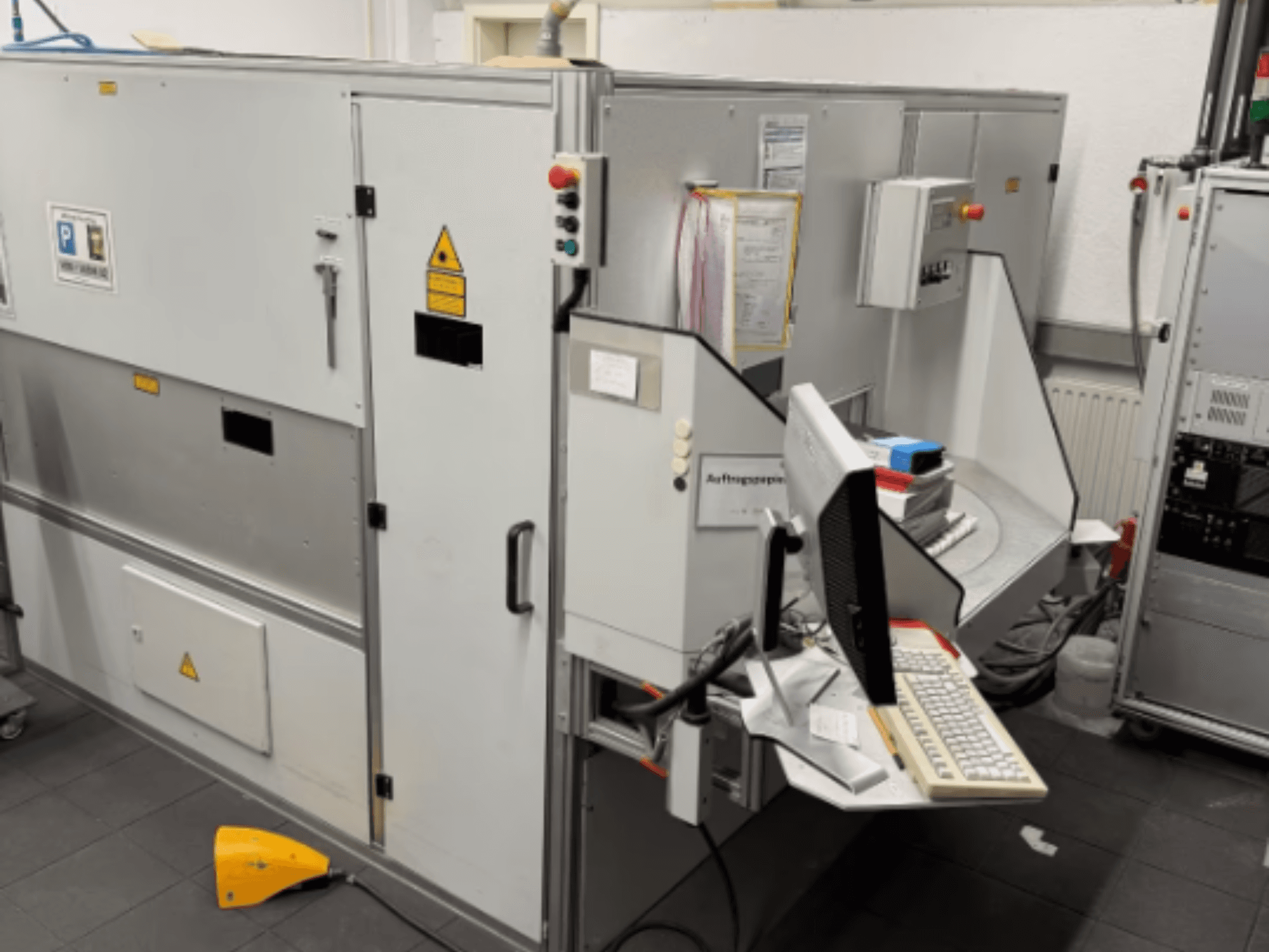 Промышленный станок с компьютерным терминалом, вид сбоку; включает кнопки управления и предупреждающие знаки безопасности.