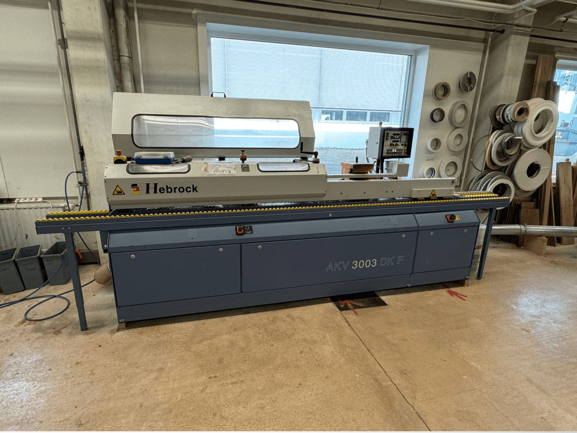 Кромкооблицовочный станок Hebrock AKV 3003 DK F, вид спереди, с конвейером и панелью управления, расположен в мастерской.