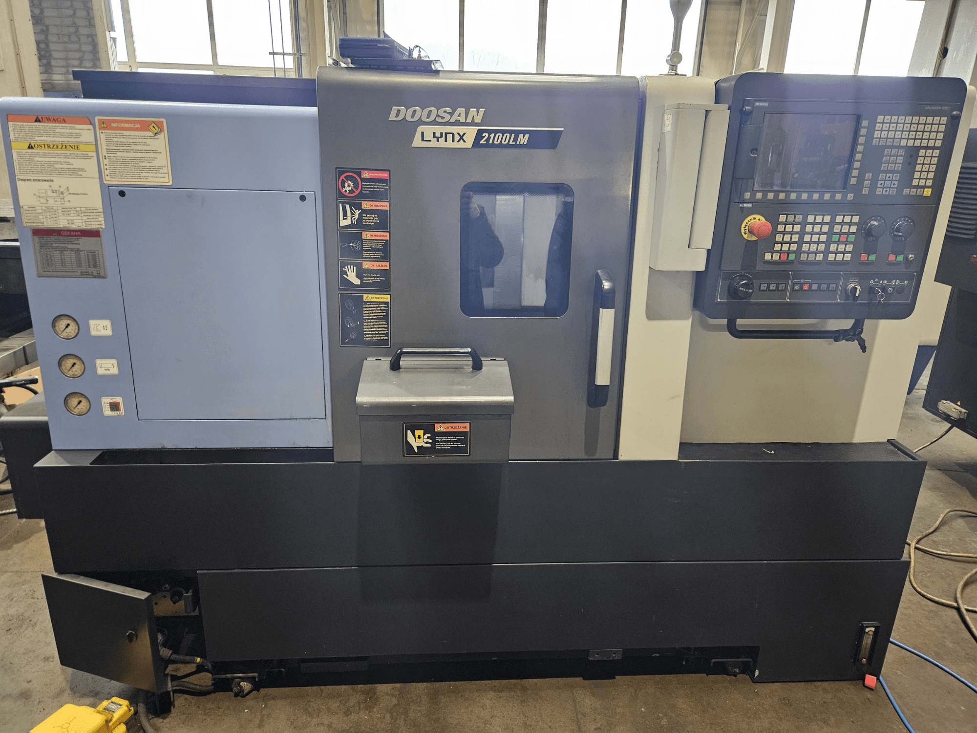 Токарный станок с ЧПУ Doosan Lynx 2100LM, вид спереди, видны панель управления, манометры давления воздуха и наклейки безопасности.