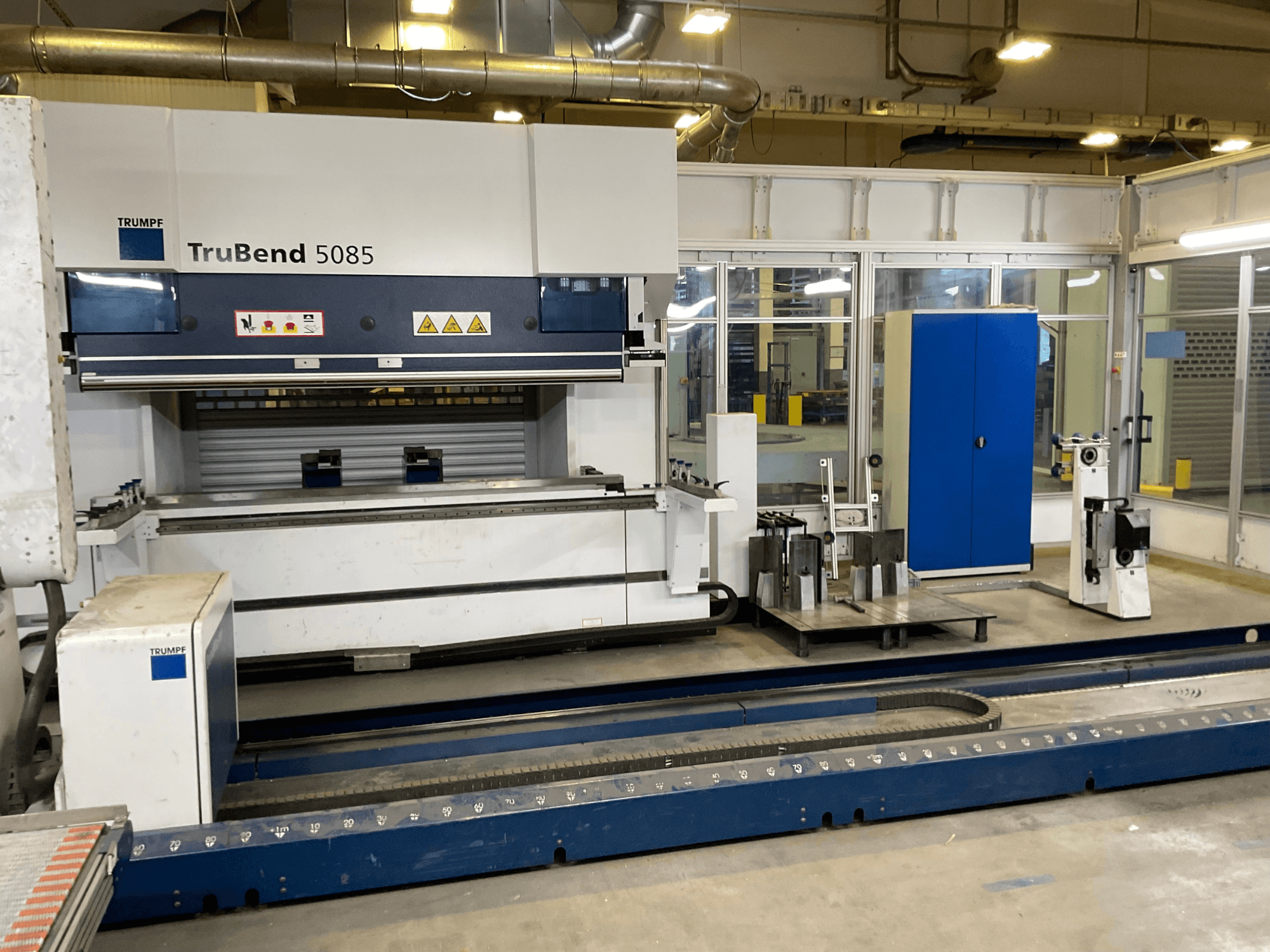 TruBend 5085 от TRUMPF, вид сбоку на большой гибочный станок для металла с панелью управления и различными инструментами рядом с ней.