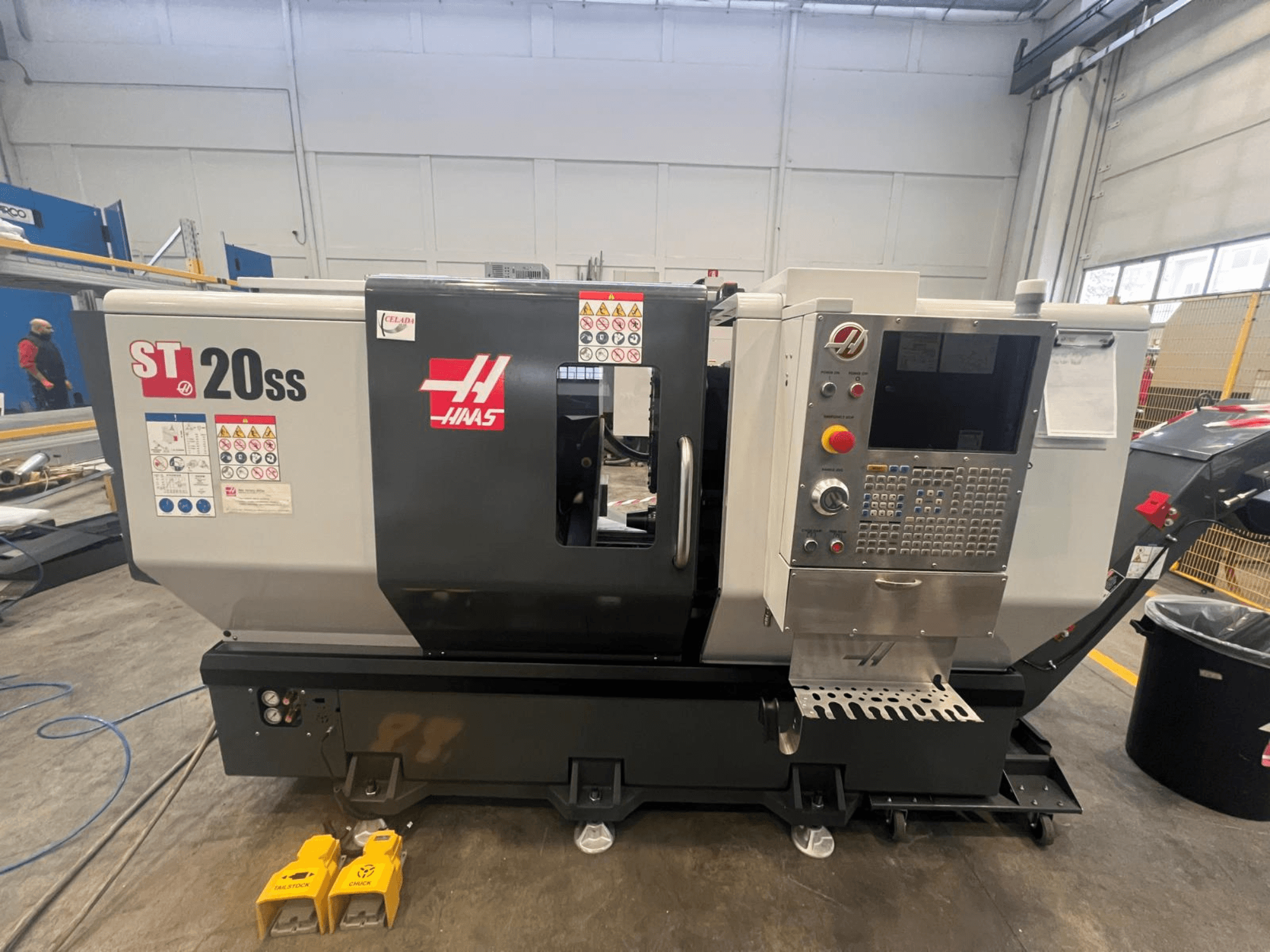 Токарный станок с ЧПУ Haas ST 20SS вид сбоку, панель управления, наклейки безопасности и ножные педали на полу.