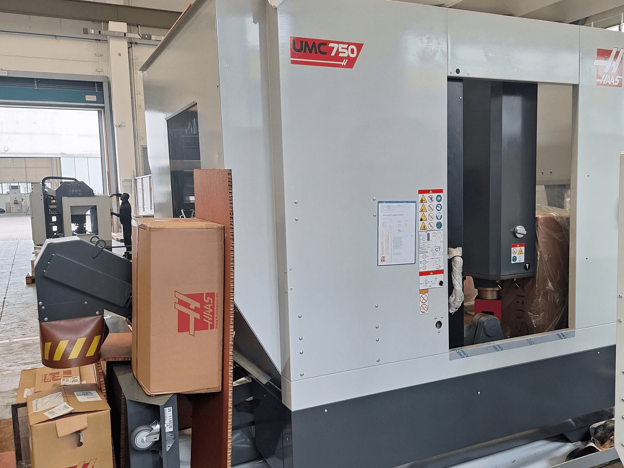 Универсальный обрабатывающий центр HAAS UMC-750, вид сбоку, на котором видны панель управления и отсек для хранения инструментов с предупреждающими надписями и упаковкой.