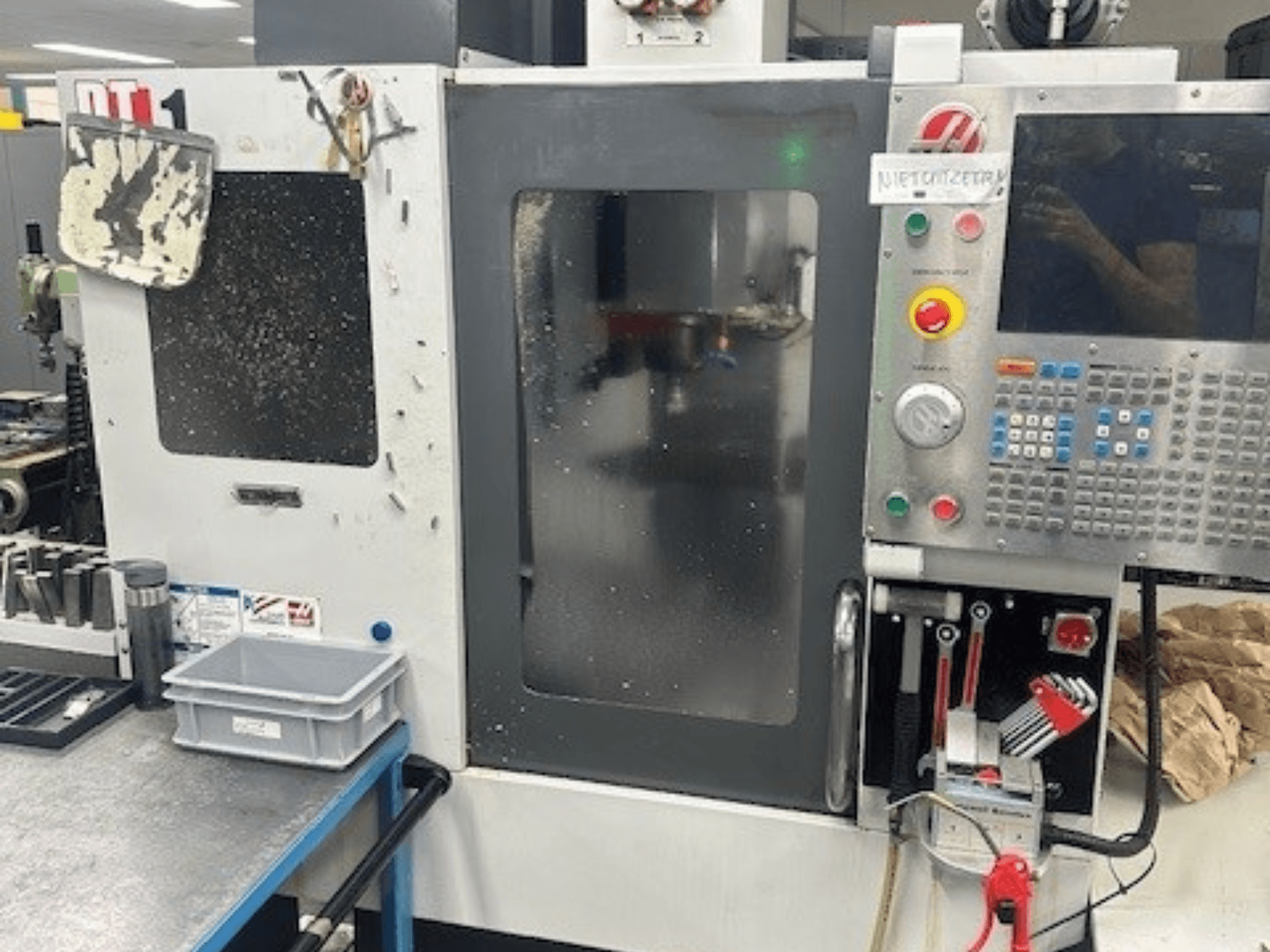 Вертикальный обрабатывающий центр Haas DT-1, вид спереди с панелью управления, видимые загрязнения и лоток для инструментов на верстаке.