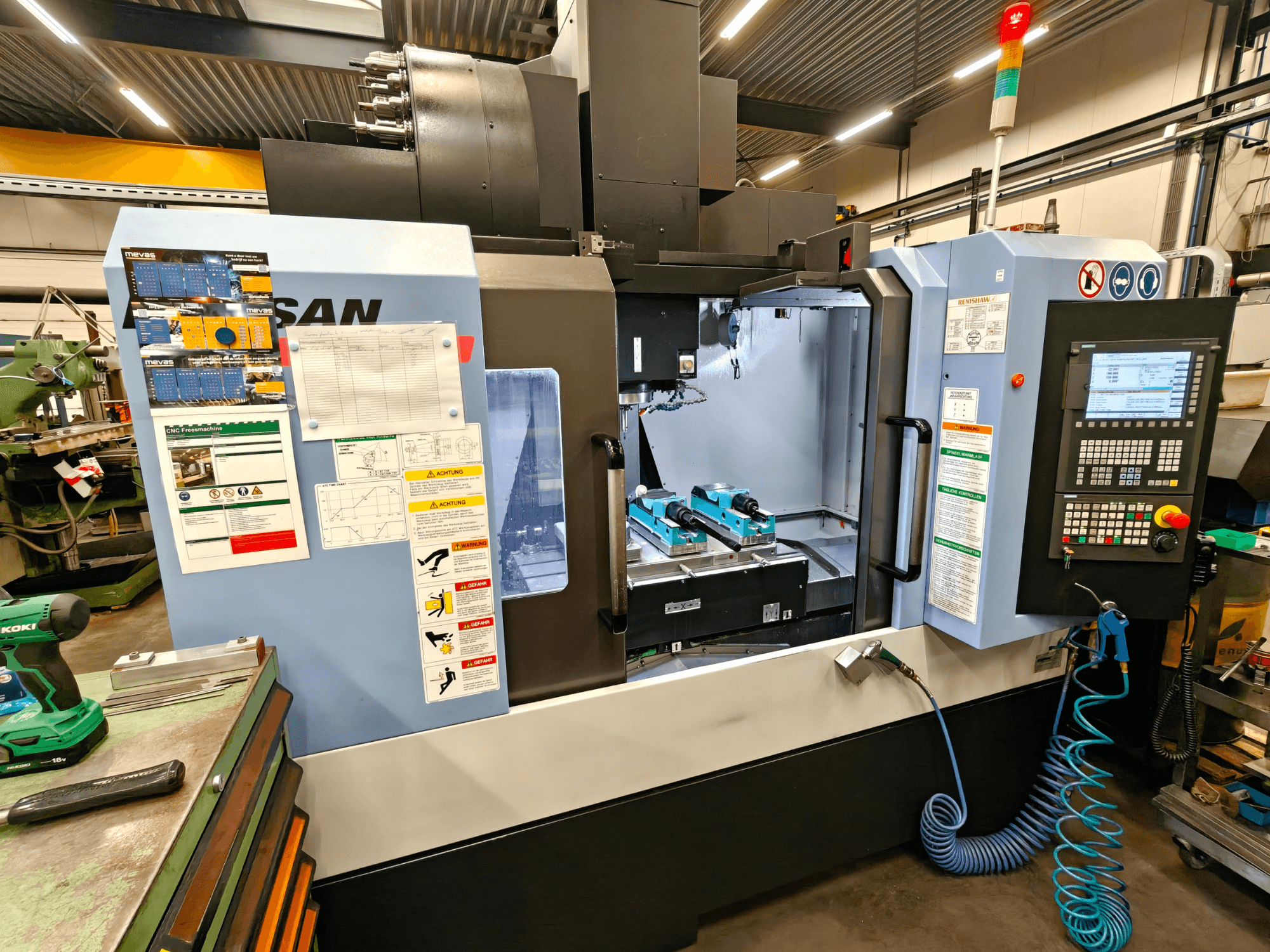 Вертикальный обрабатывающий центр DOOSAN DNM 400 II в мастерской, с элементами управления, держателями инструментов и знаками безопасности, вид спереди.