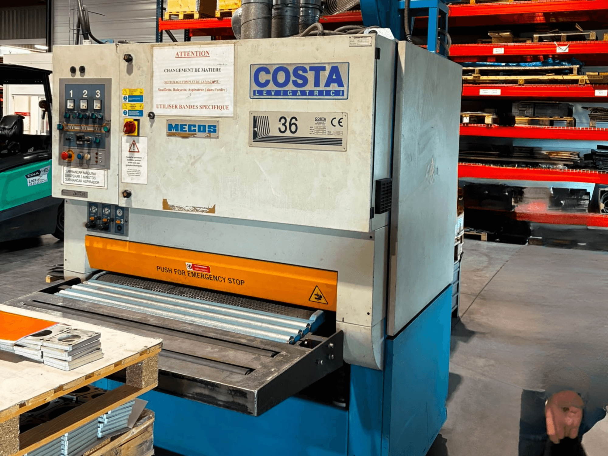 Машина для удаления заусенцев Costa M36 CCS 1150 в виде сбоку, с панелью управления, кнопкой аварийной остановки и конвейерной поверхностью.