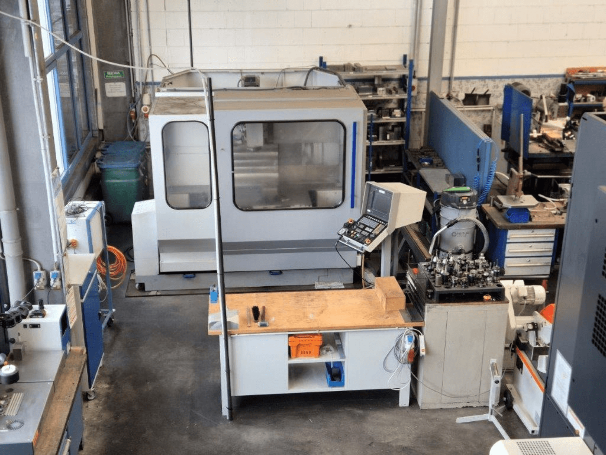 Фрезерный станок с ЧПУ в мастерской, вид спереди, на заднем плане видны панель управления, рабочая поверхность и инструменты.