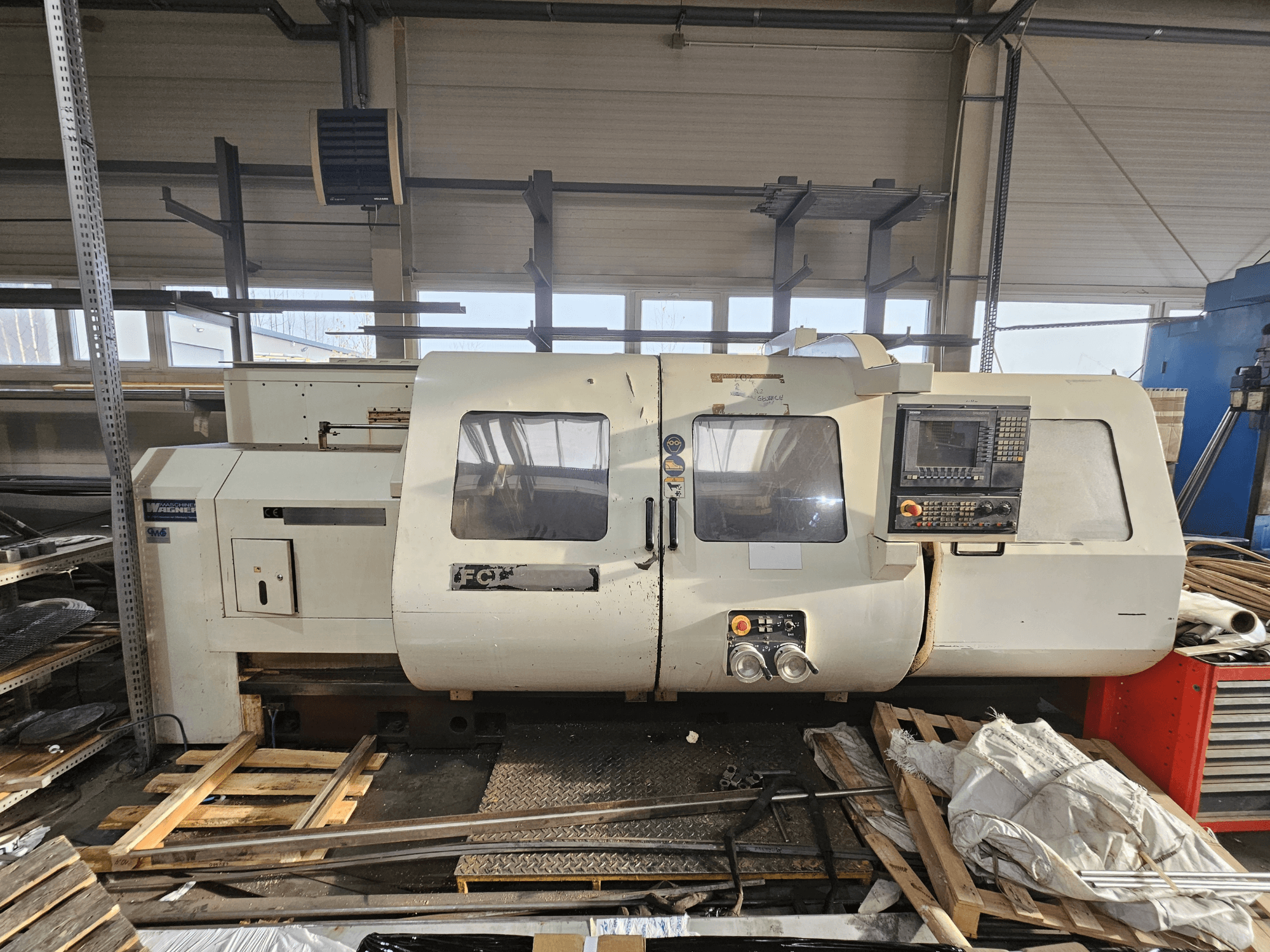 Токарный станок с ЧПУ Wabeco, вид сбоку, панель управления, окна и рабочая зона; станки на деревянных поддонах в мастерской.